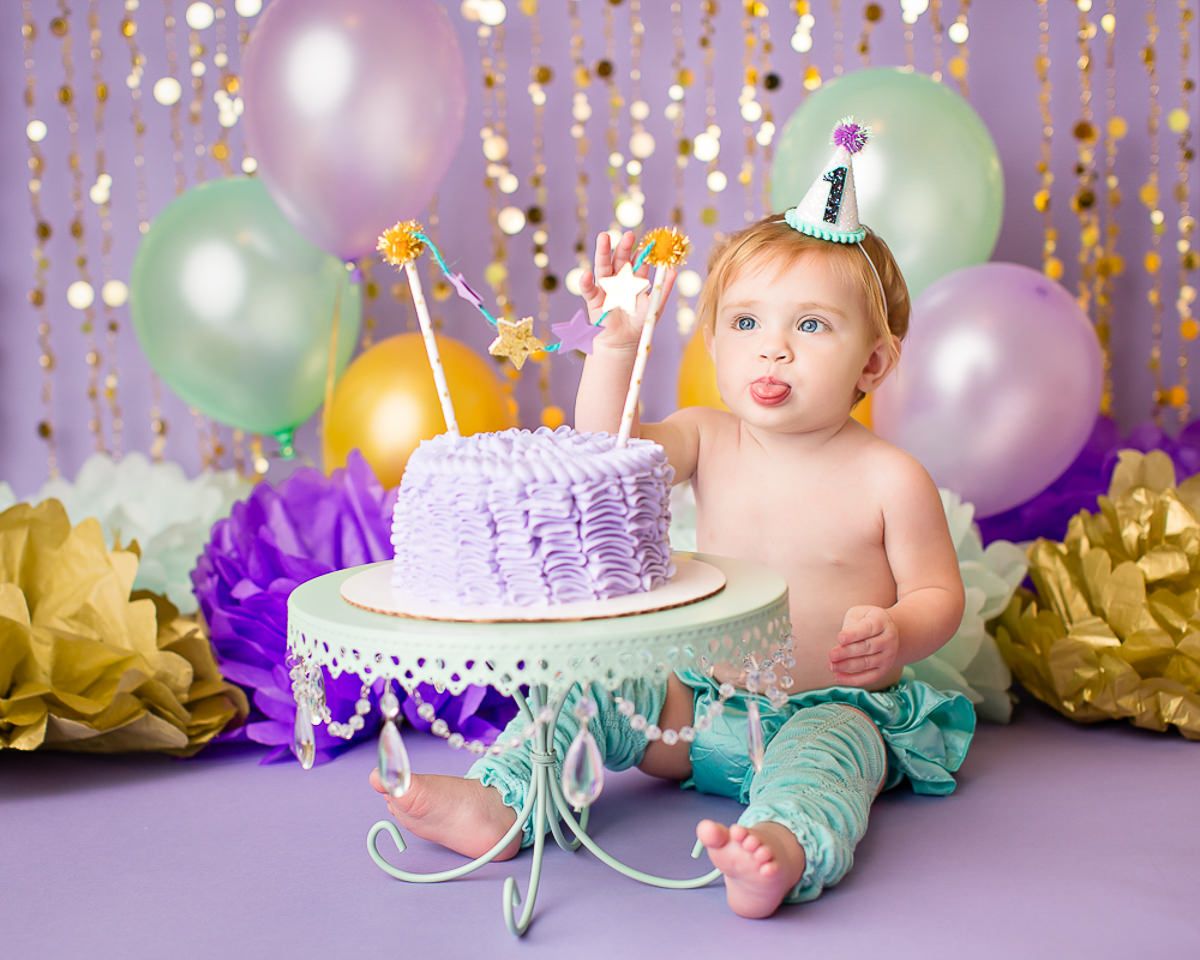 Baby girl in teal leg warmers celebrating first birthday smashing purple ruffle cake with balloons and party hat.