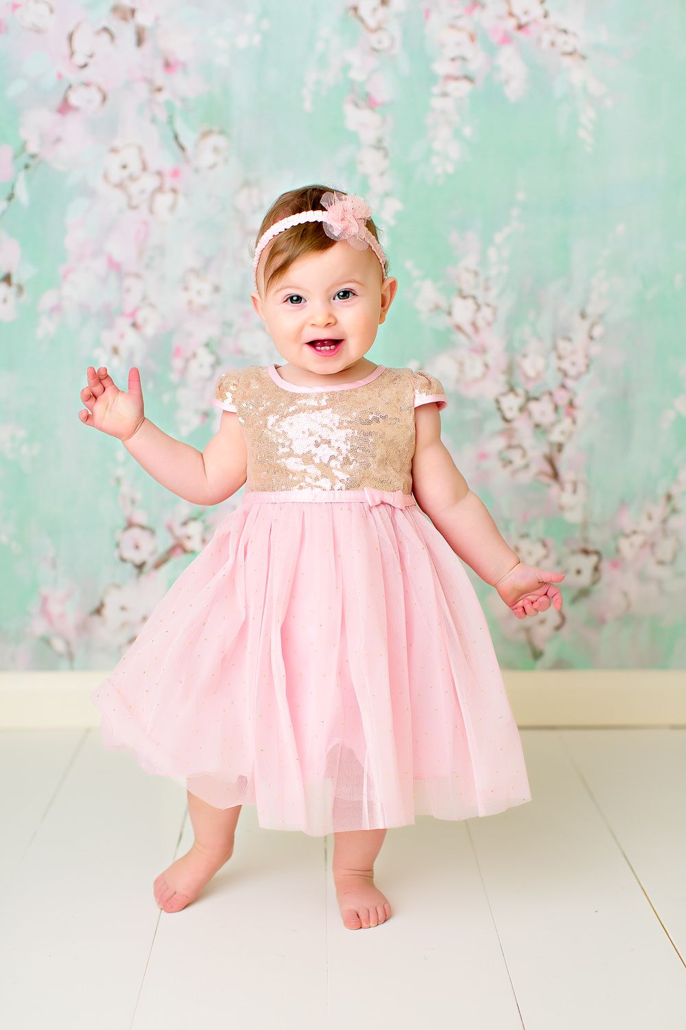Adorable baby girl in gold sequin and pink tulle dress standing before a floral mint backdrop.