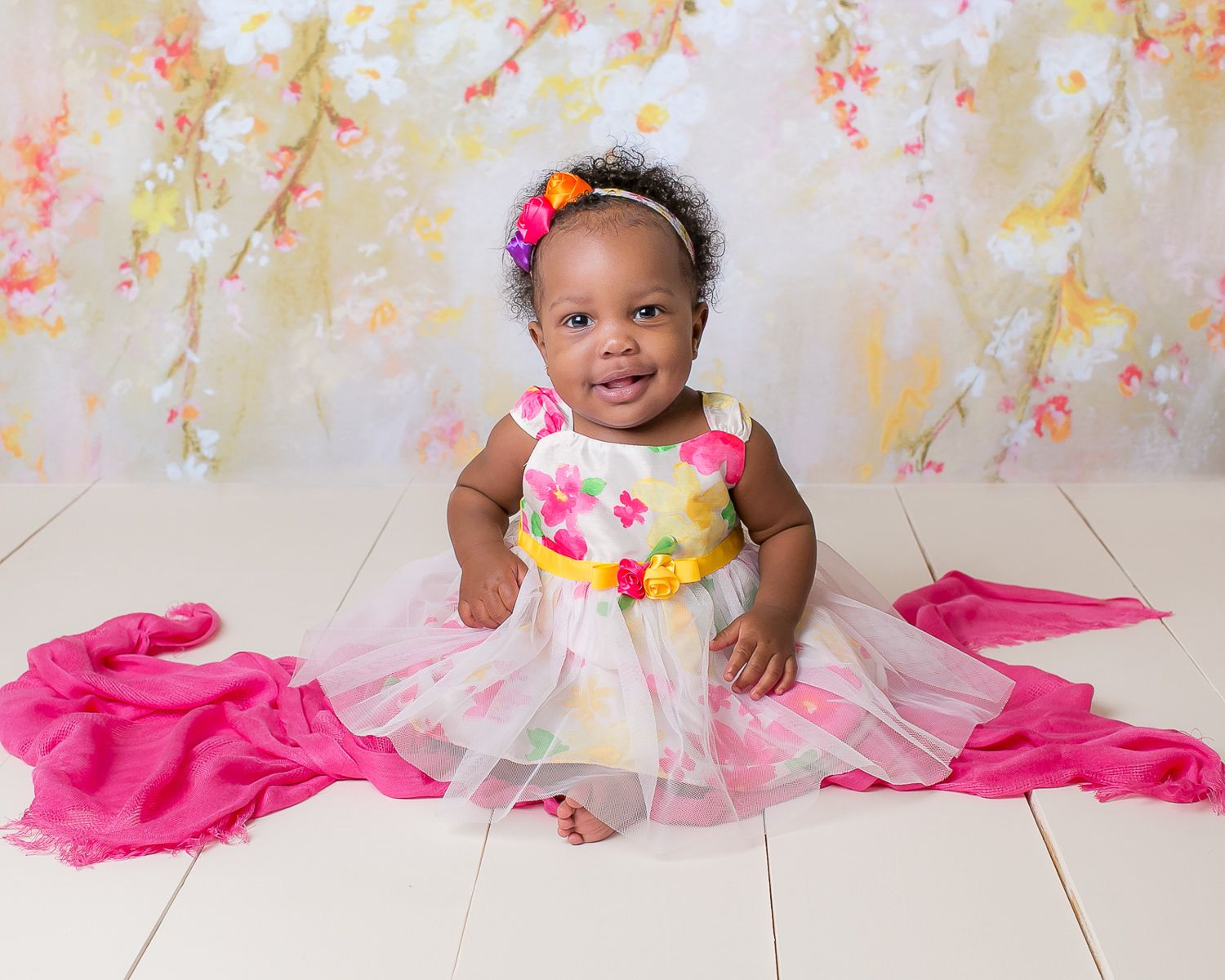 Happy baby girl in colorful floral dress sitting on white floor with pink fabric backdrop.