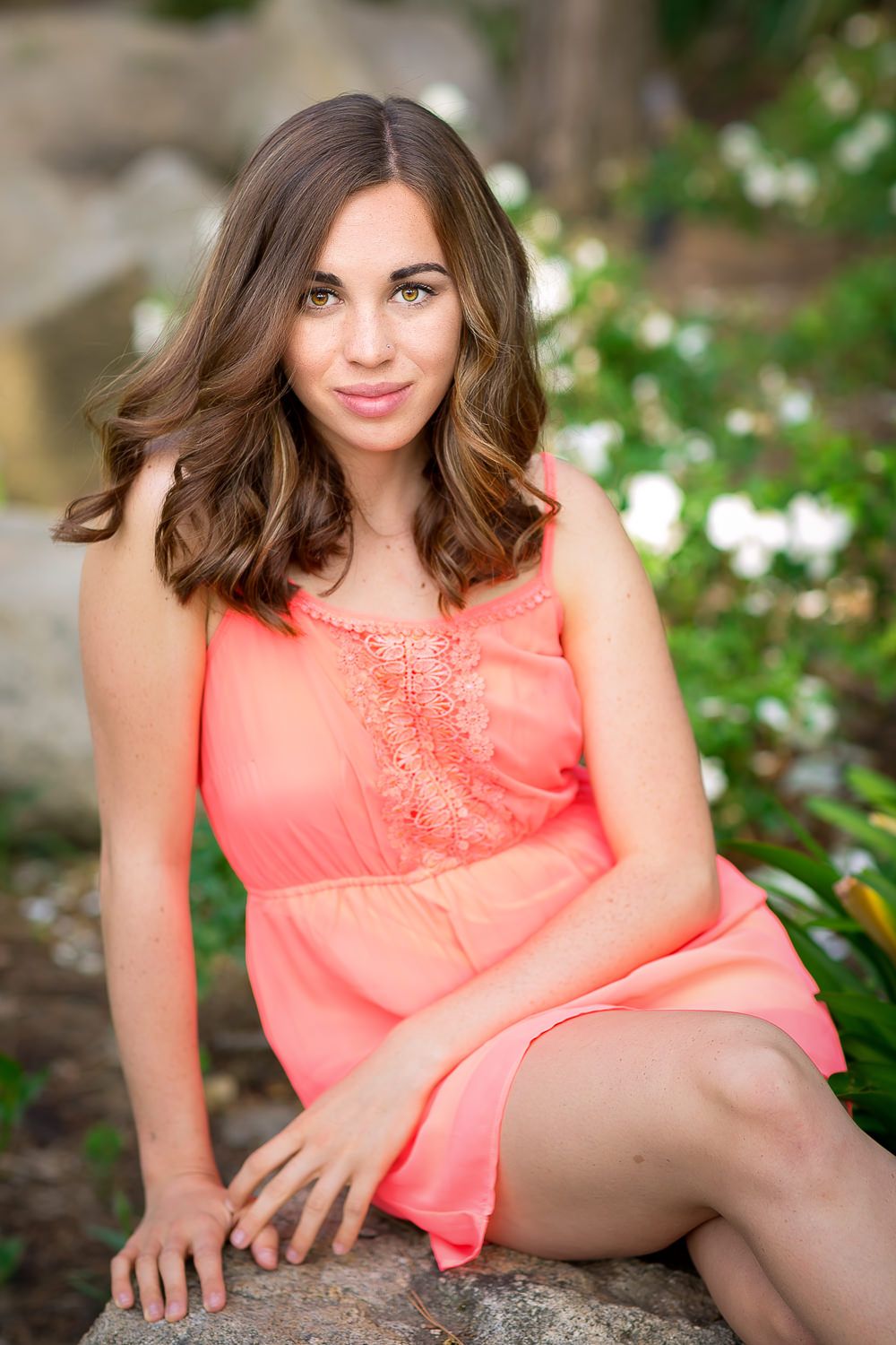Young woman in coral dress sitting on rocks in a garden with white flowers and greenery.