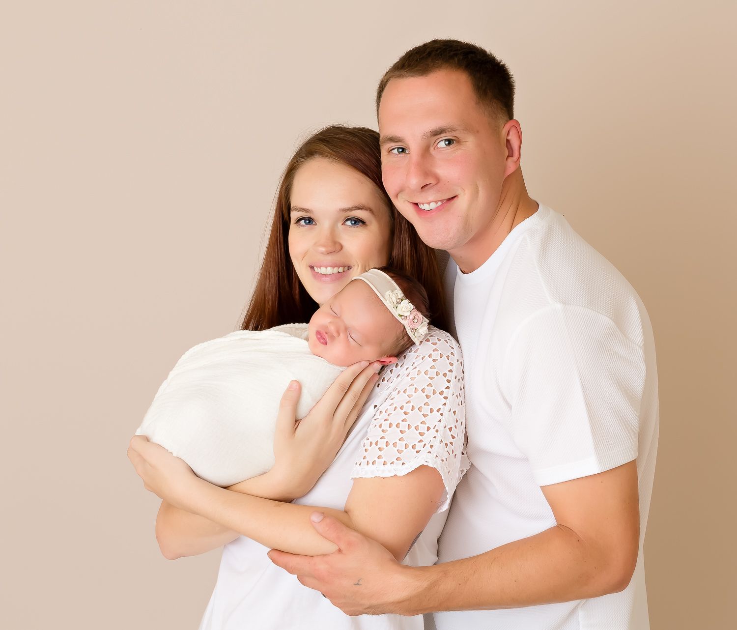 Smiling young couple in white shirts proudly holds their newborn baby in white wrap.