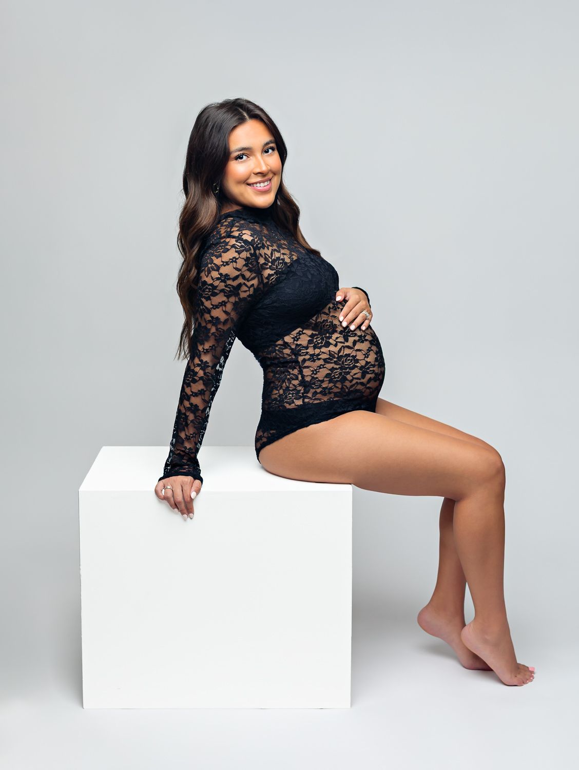 Pregnant woman in black lace bodysuit poses gracefully on white cube for a professional maternity photo shoot.