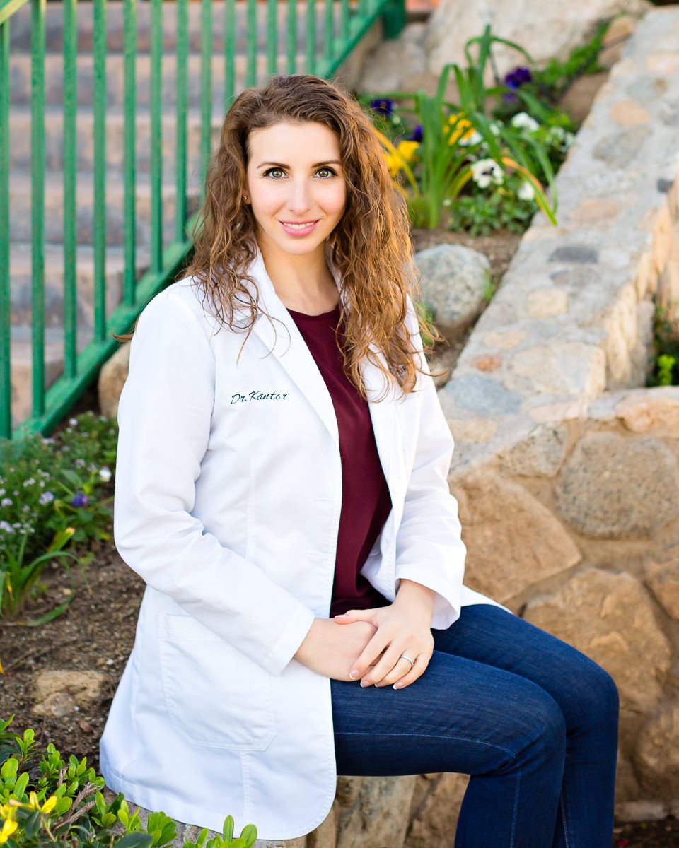 Female doctor in white lab coat sitting outdoors near rock garden and green fence, smiling professionally.
