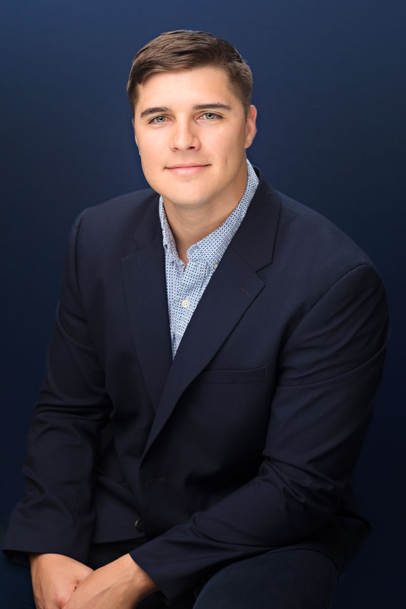 Professional headshot of a young man in a navy blazer and checkered shirt, smiling confidently against a dark blue background.