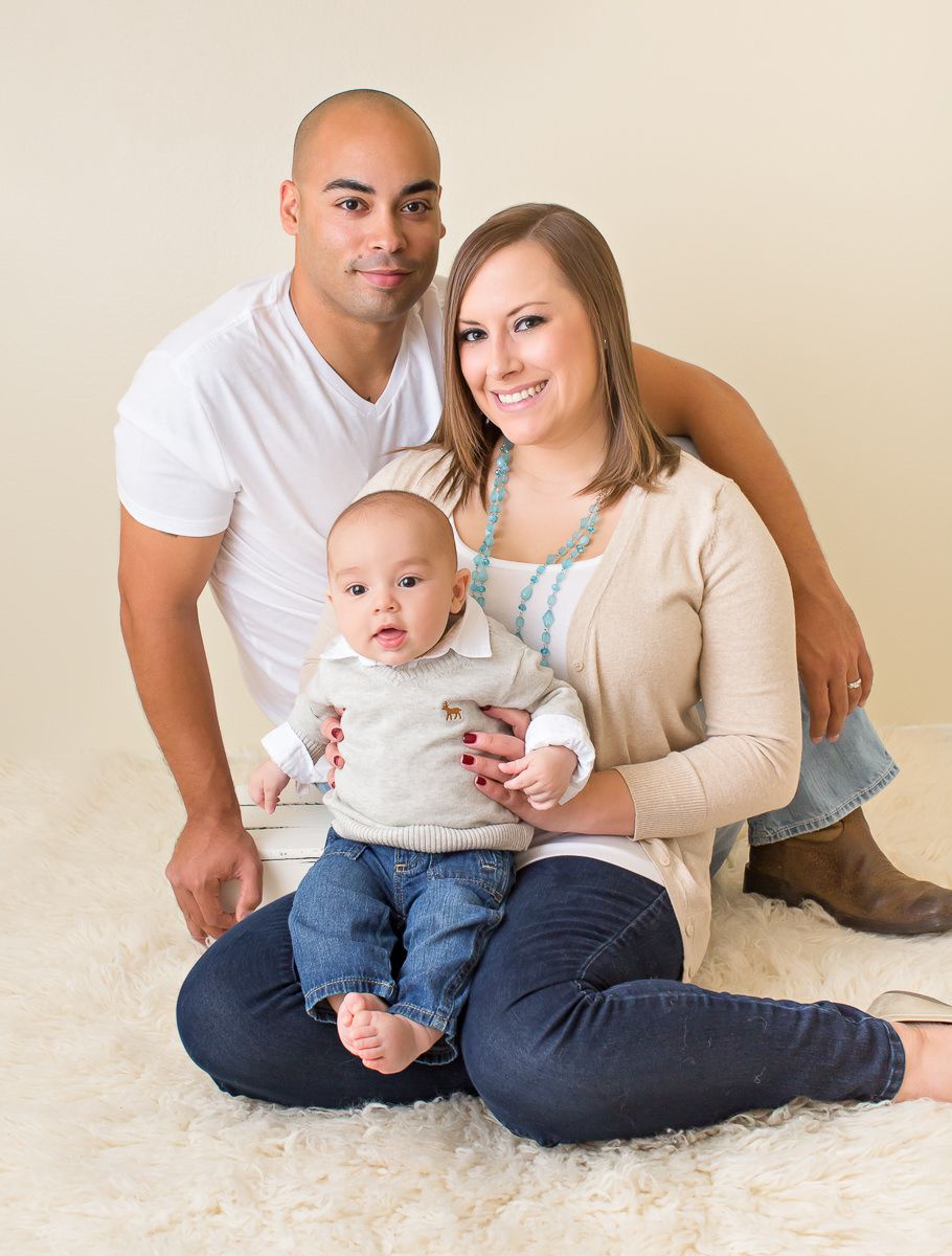 Young family portrait with bald father, smiling mother, and cute baby boy on fur rug.