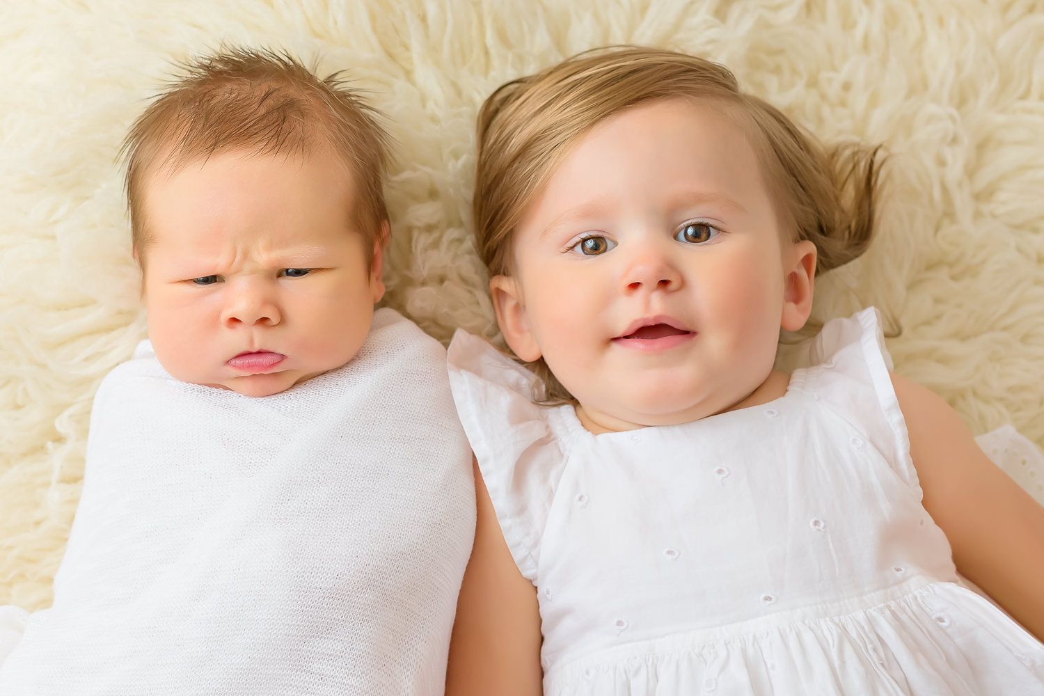 Two adorable babies lying on a fluffy white blanket, one frowning and one smiling with blue eyes.
