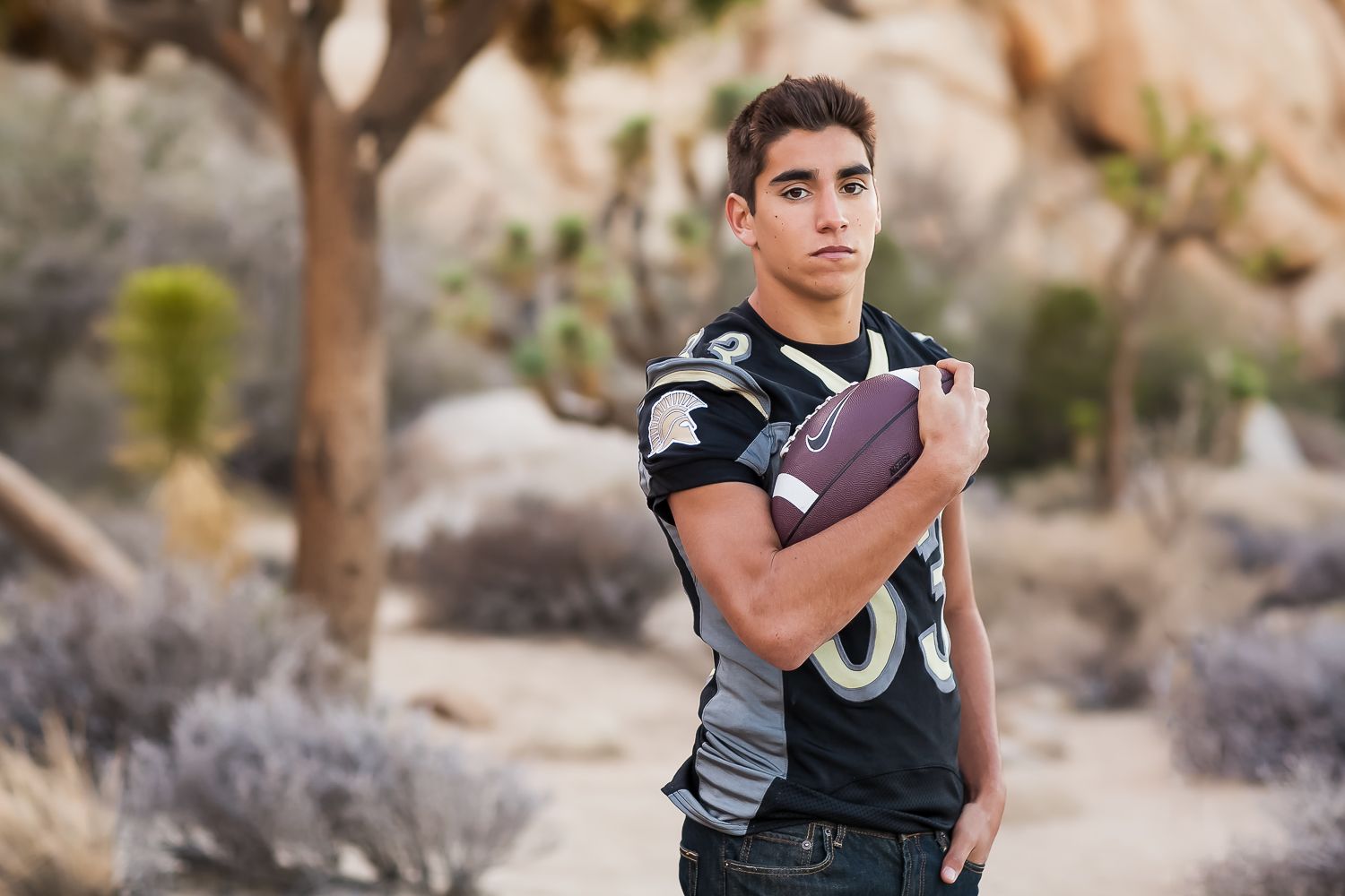 Teen boy in black football jersey holding a football outdoors in a desert landscape with Joshua trees.
