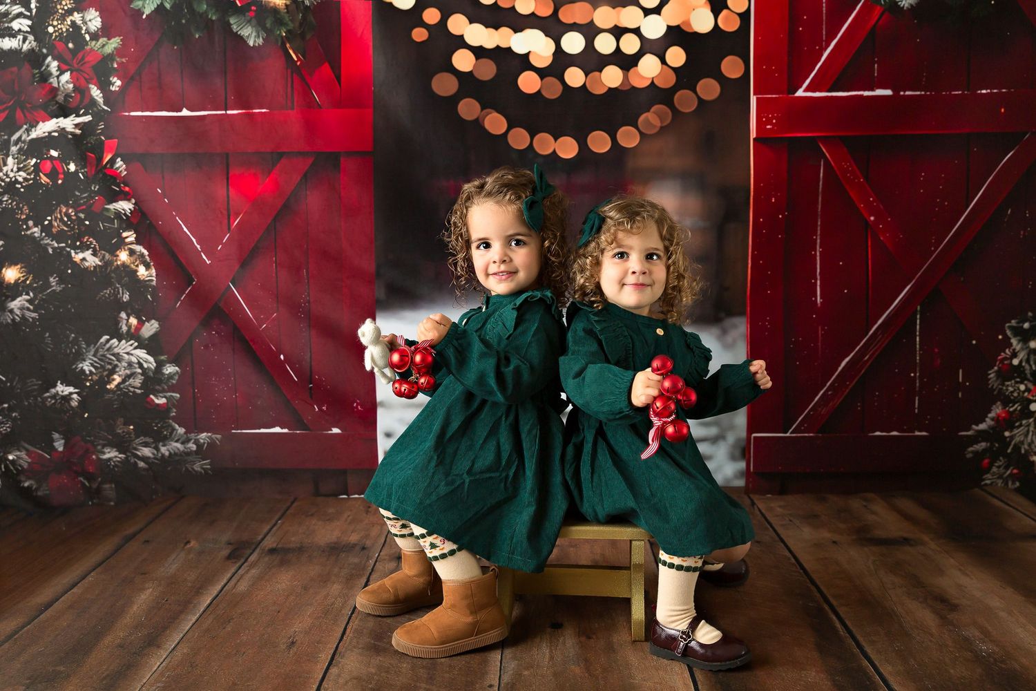 Two young girls in matching green dresses hold red ornaments beside a Christmas tree and red barn doors.