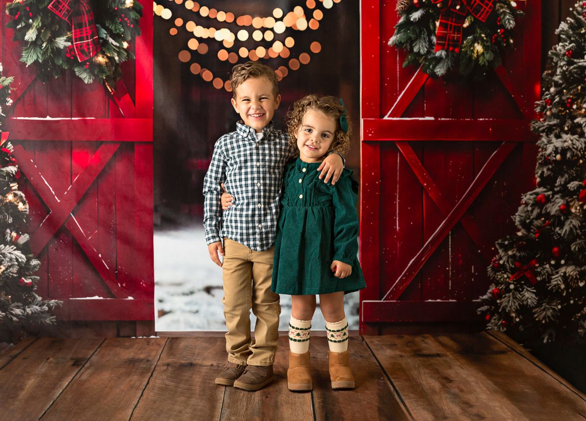 Two children pose in front of red barn doors with Christmas decorations, boy in plaid shirt and girl in green dress.
