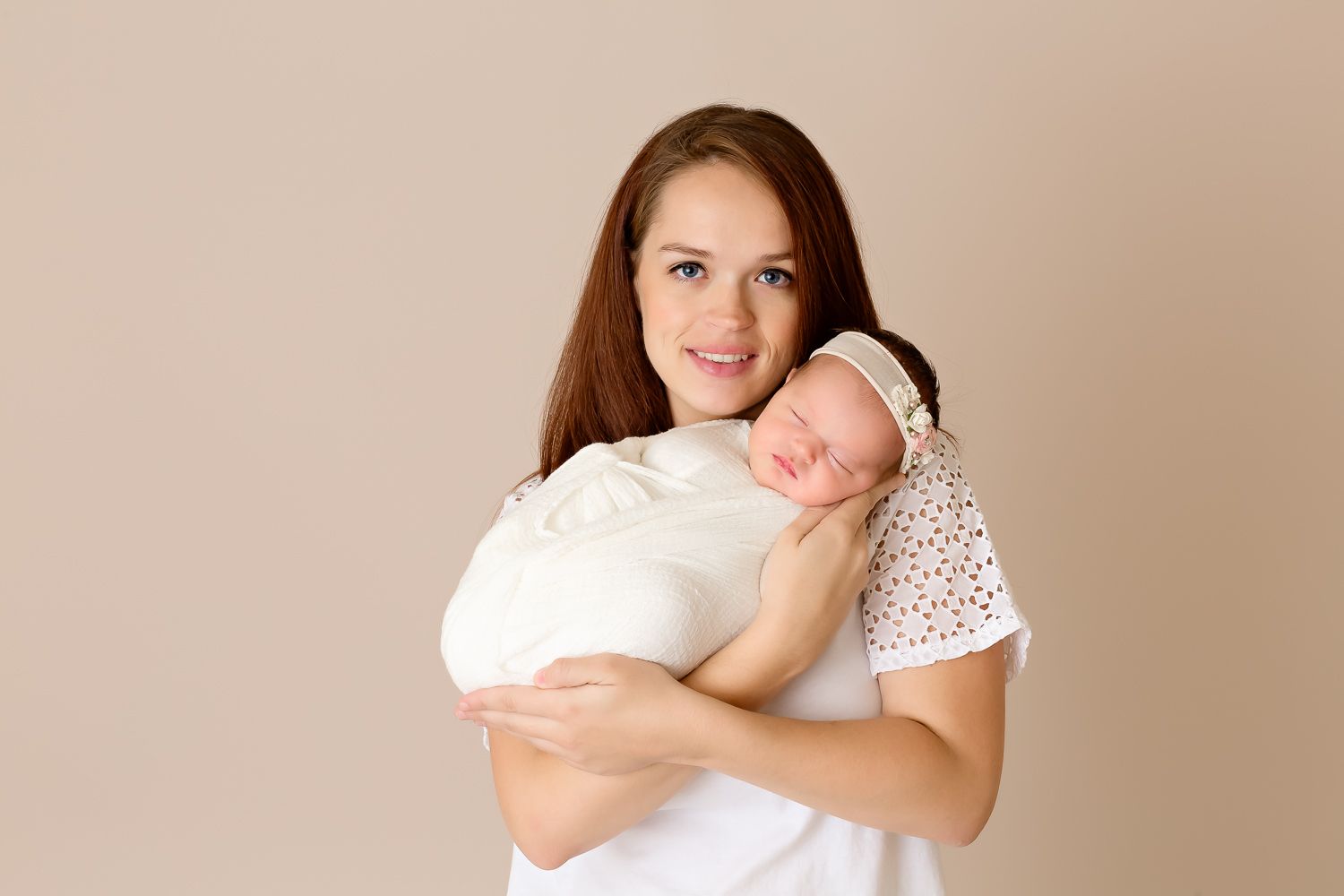 Smiling brunette mother holding her sleeping swaddled newborn baby against her shoulder on beige background.