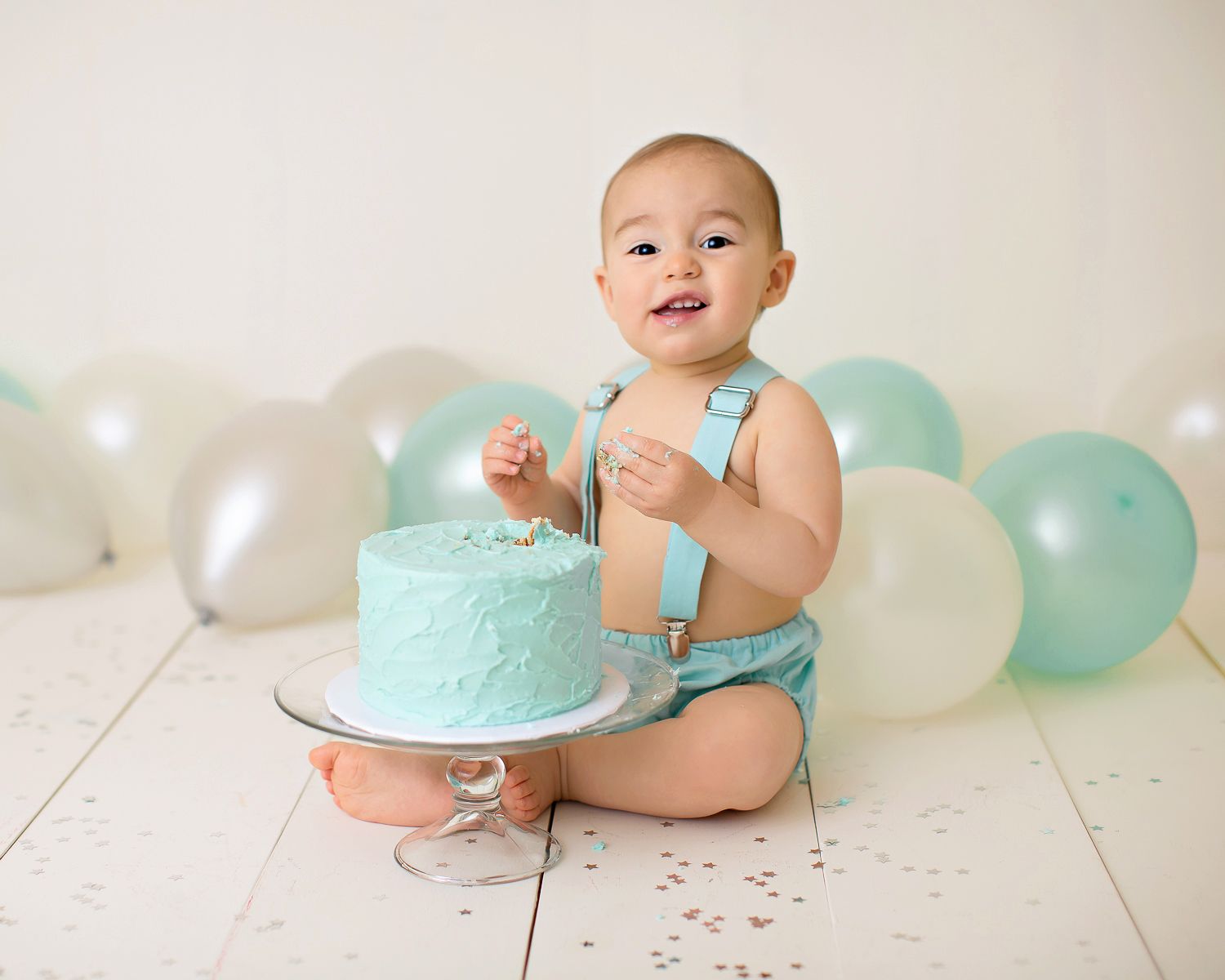 Baby boy in blue suspenders smashes first birthday cake surrounded by mint and white balloons on white floor.