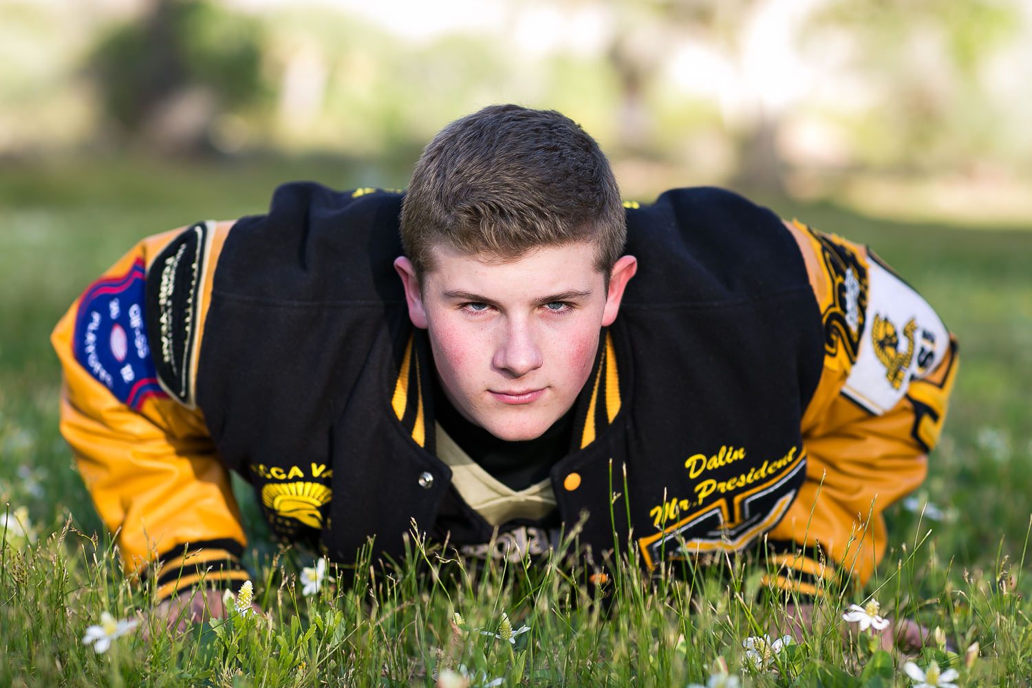 Young man in black and gold letterman jacket posing in grass for a sports senior portrait photo.