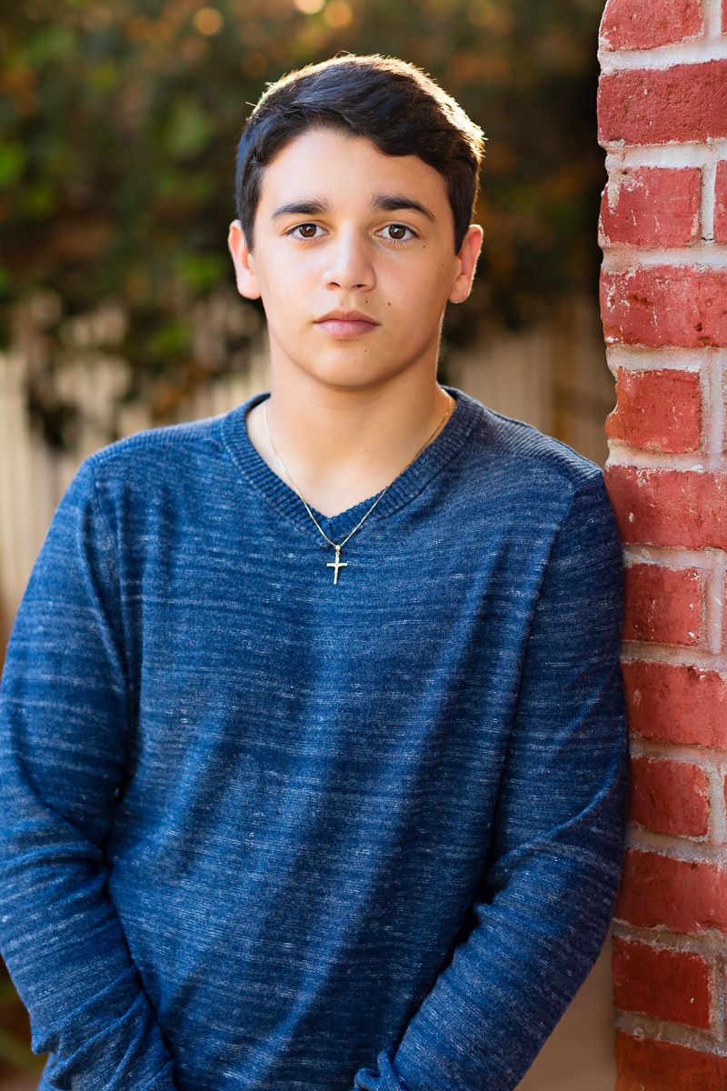 Teen boy in blue sweater with gold cross necklace leaning against red brick wall outdoors.