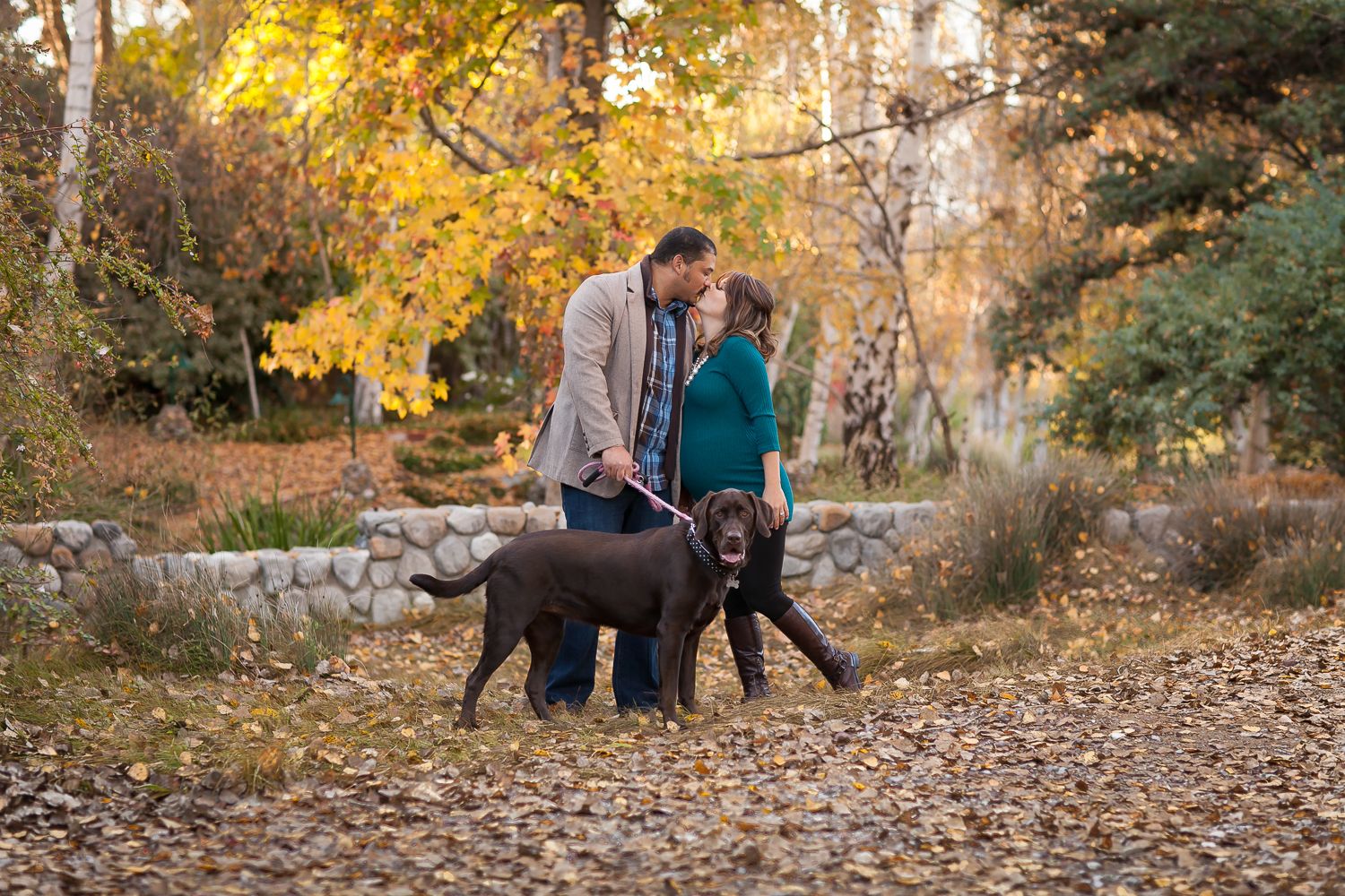 Couple sharing a romantic moment with their chocolate Labrador on a leaf-covered path surrounded by autumn foliage.