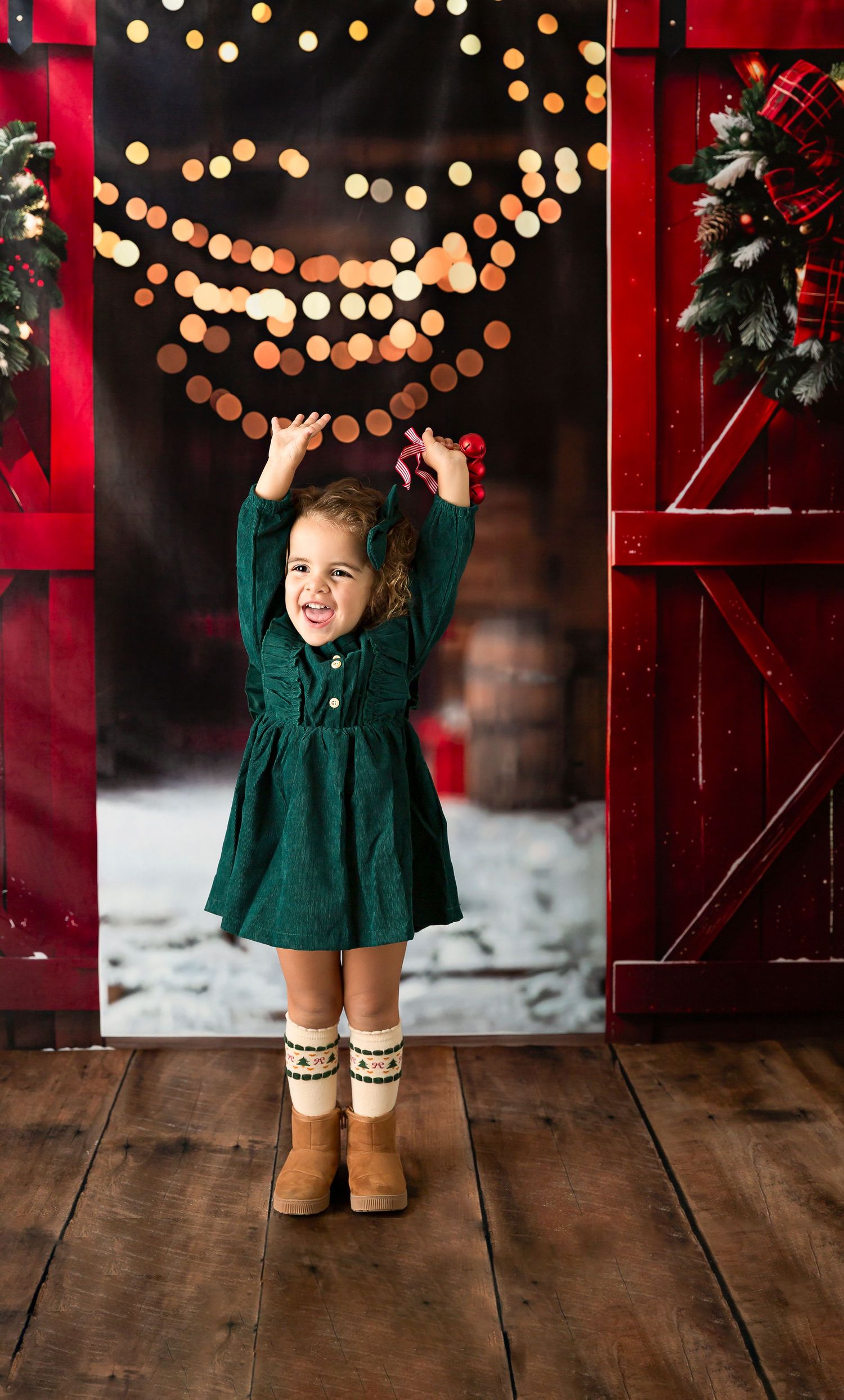 A joyful toddler in a green dress and UGG boots poses before a festive Christmas backdrop with bokeh lights.
