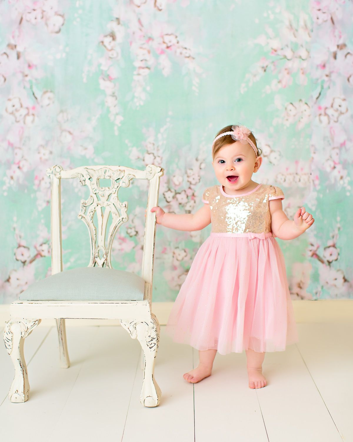 Smiling baby girl in pink gold dress standing beside vintage chair on floral backdrop.