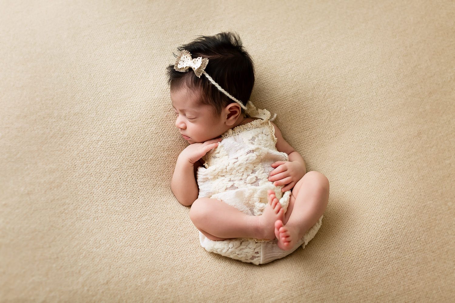 Peaceful newborn baby girl in cream lace romper with bow headband on beige backdrop.