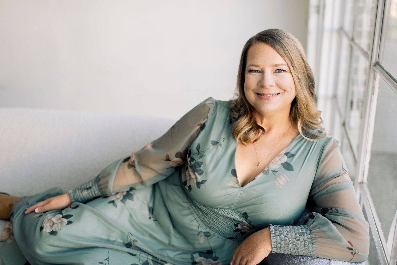 Woman in floral sage green dress smiling on white sofa near bright window in elegant portrait photo.