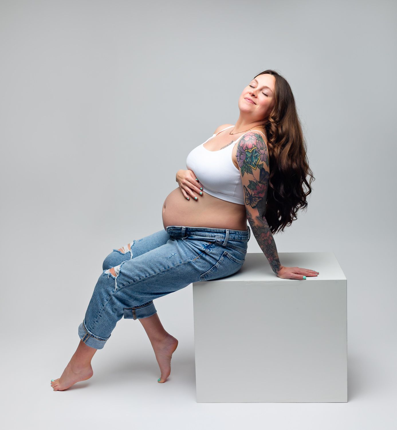 Tattooed pregnant woman in white crop top and jeans relaxing on white cube on gray background.