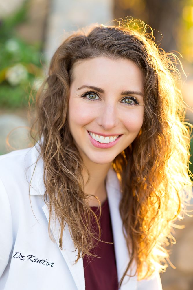 Dr. Kantor smiling in a white lab coat with curly brown hair and green eyes outdoors in warm sunlight.
