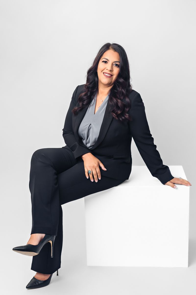 Professional woman in black business suit sitting on white cube, smiling confidently against light gray background.