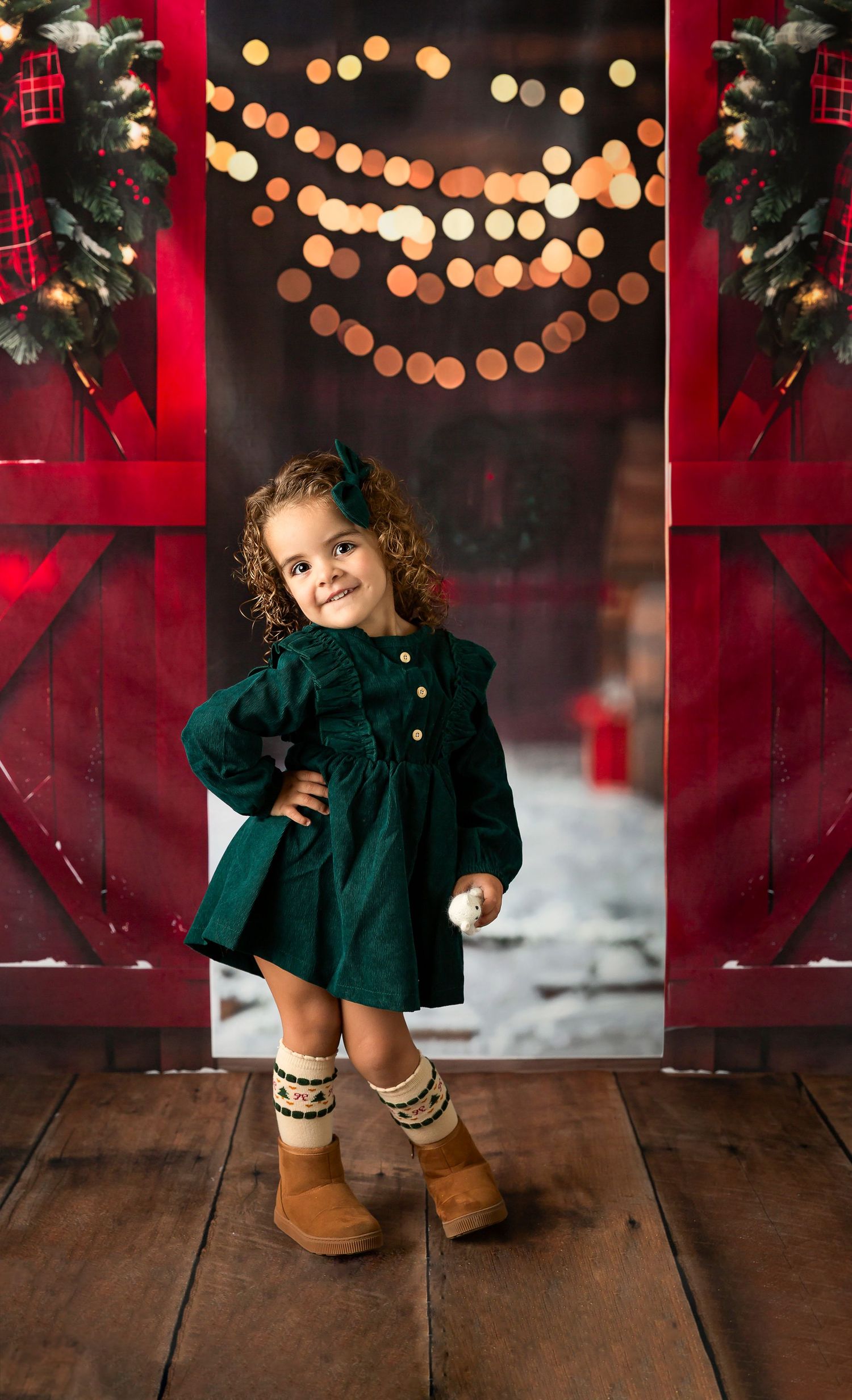 A smiling toddler girl in a green dress and UGG boots poses in a festive red barn doorway with Christmas lights.