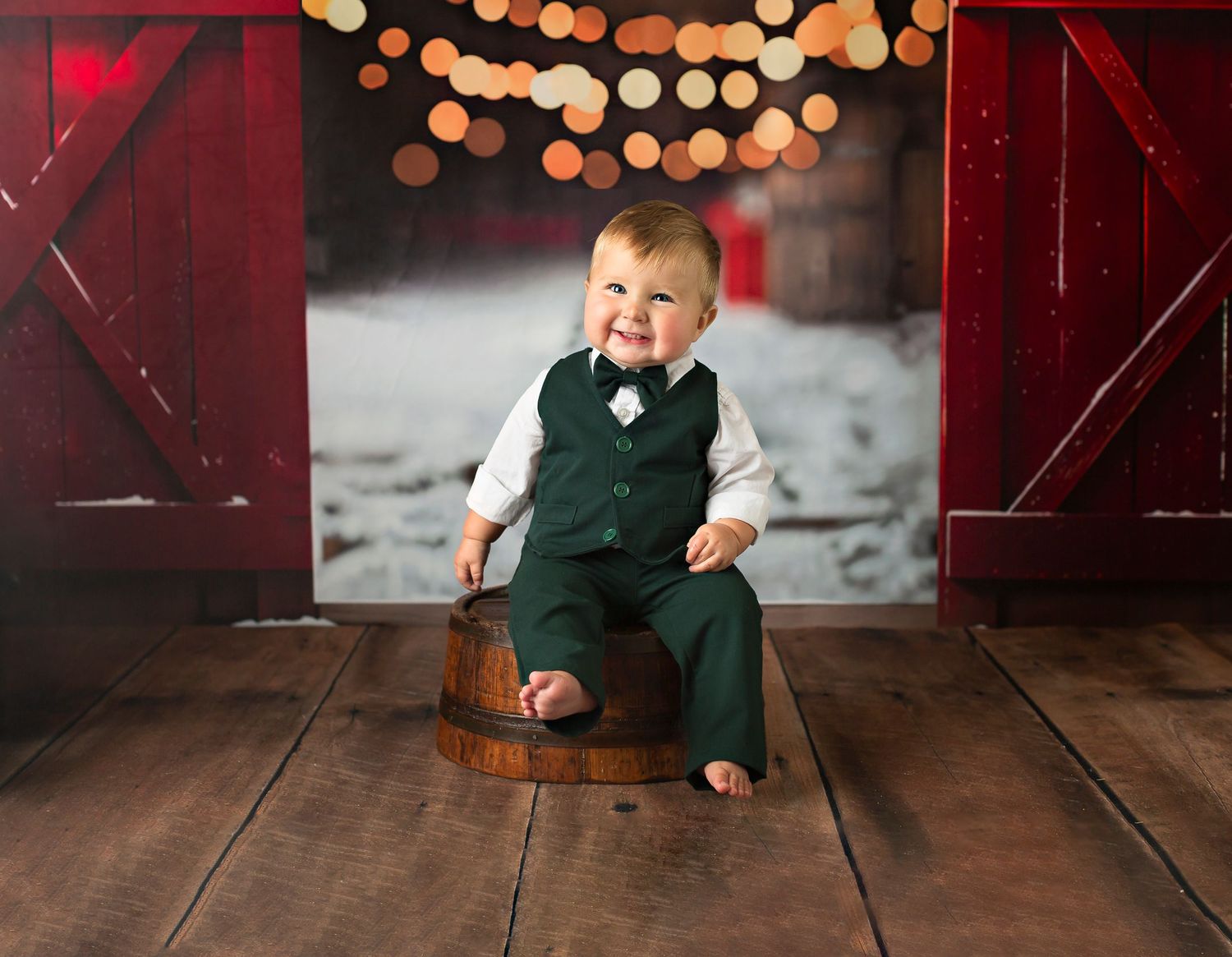 Smiling baby boy in green vest suit sitting on wooden barrel with festive bokeh lights and red barn doors backdrop.