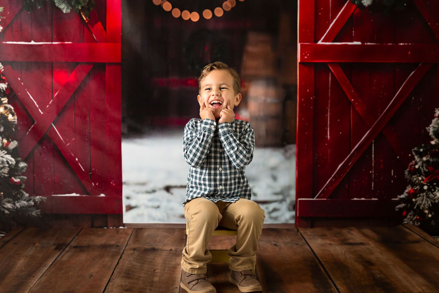 A joyful young boy sits between red barn doors with Christmas trees, wearing a plaid shirt and khaki pants.