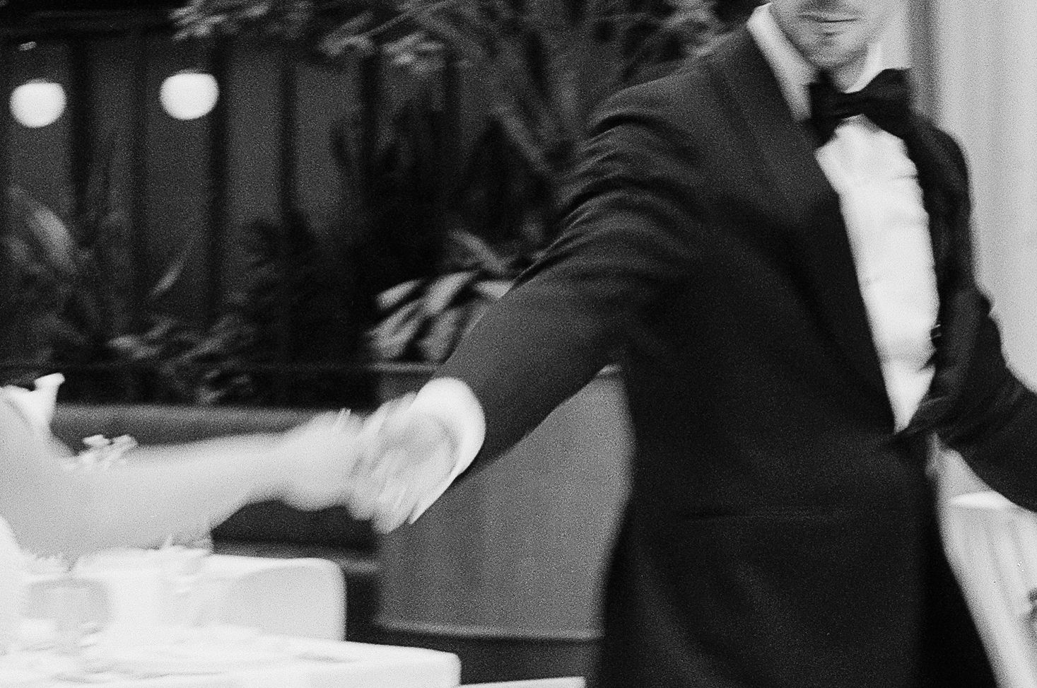 Groom in black tuxedo and bow tie dancing and holding hands with bride at elegant reception in black and white, grainy film photo.