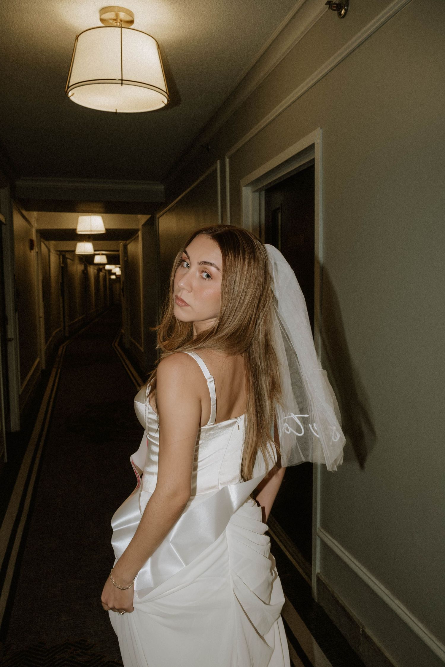 A bride in a white wedding dress and veil poses dramatically in a dimly lit hotel hallway.