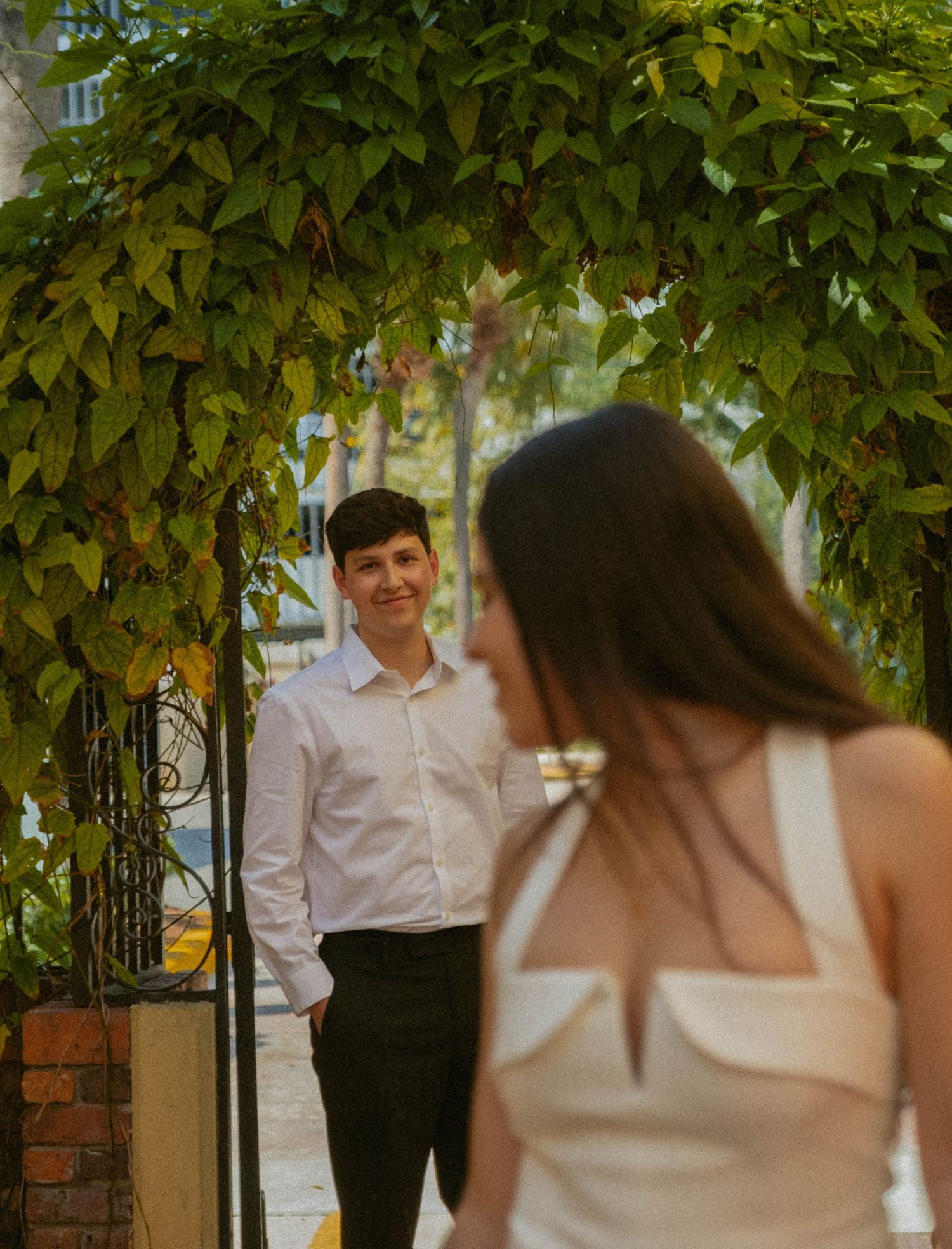 A couple shares a romantic moment under a lush green pergola in an outdoor garden setting.