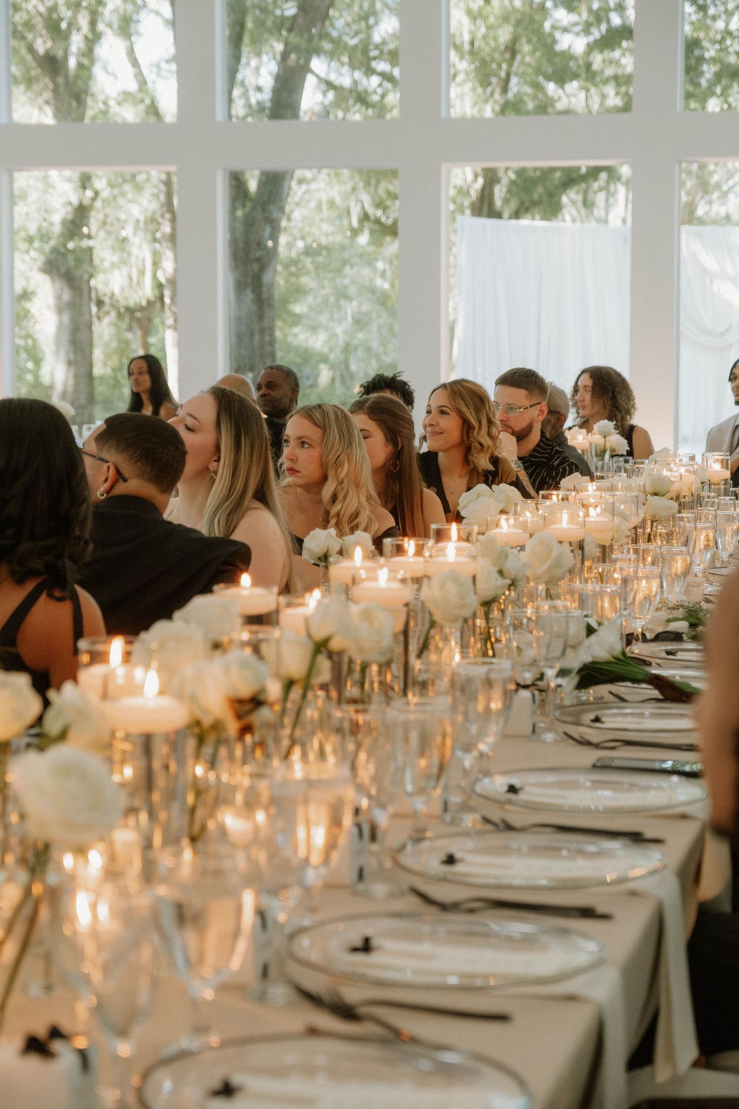 Elegant wedding reception table setting with glowing candles and white linens stretches into the distance at an indoor venue.