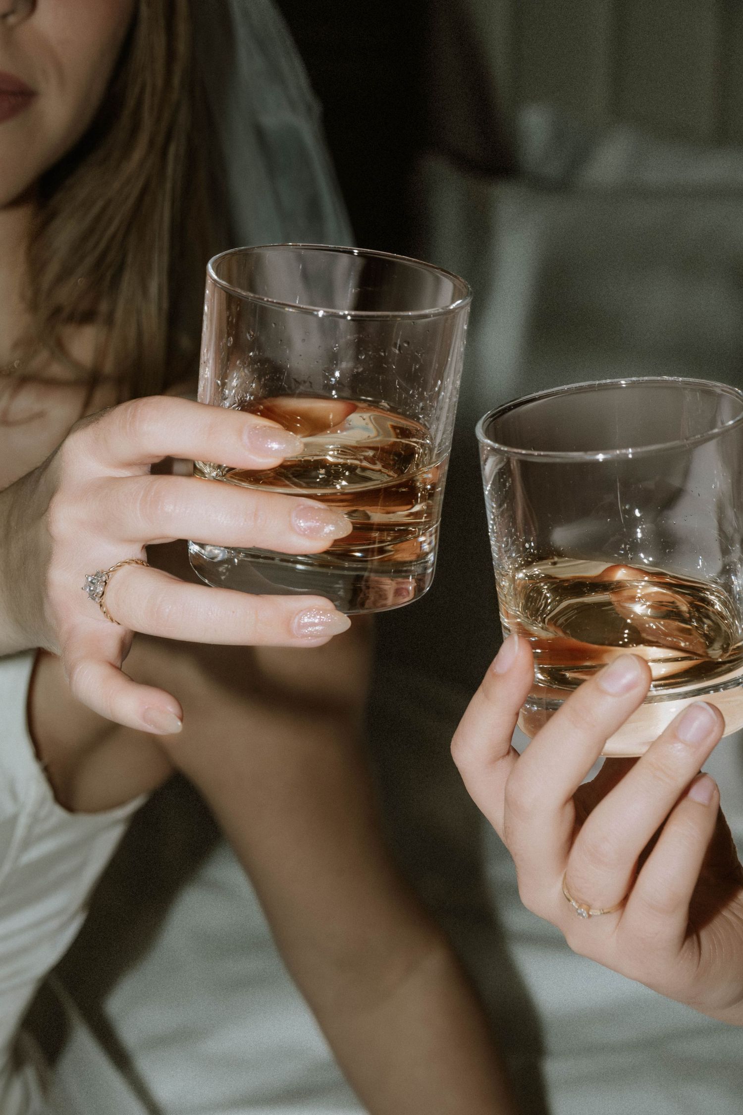 Two hands holding glasses of whiskey or bourbon with ice for a celebratory toast in moody lighting.