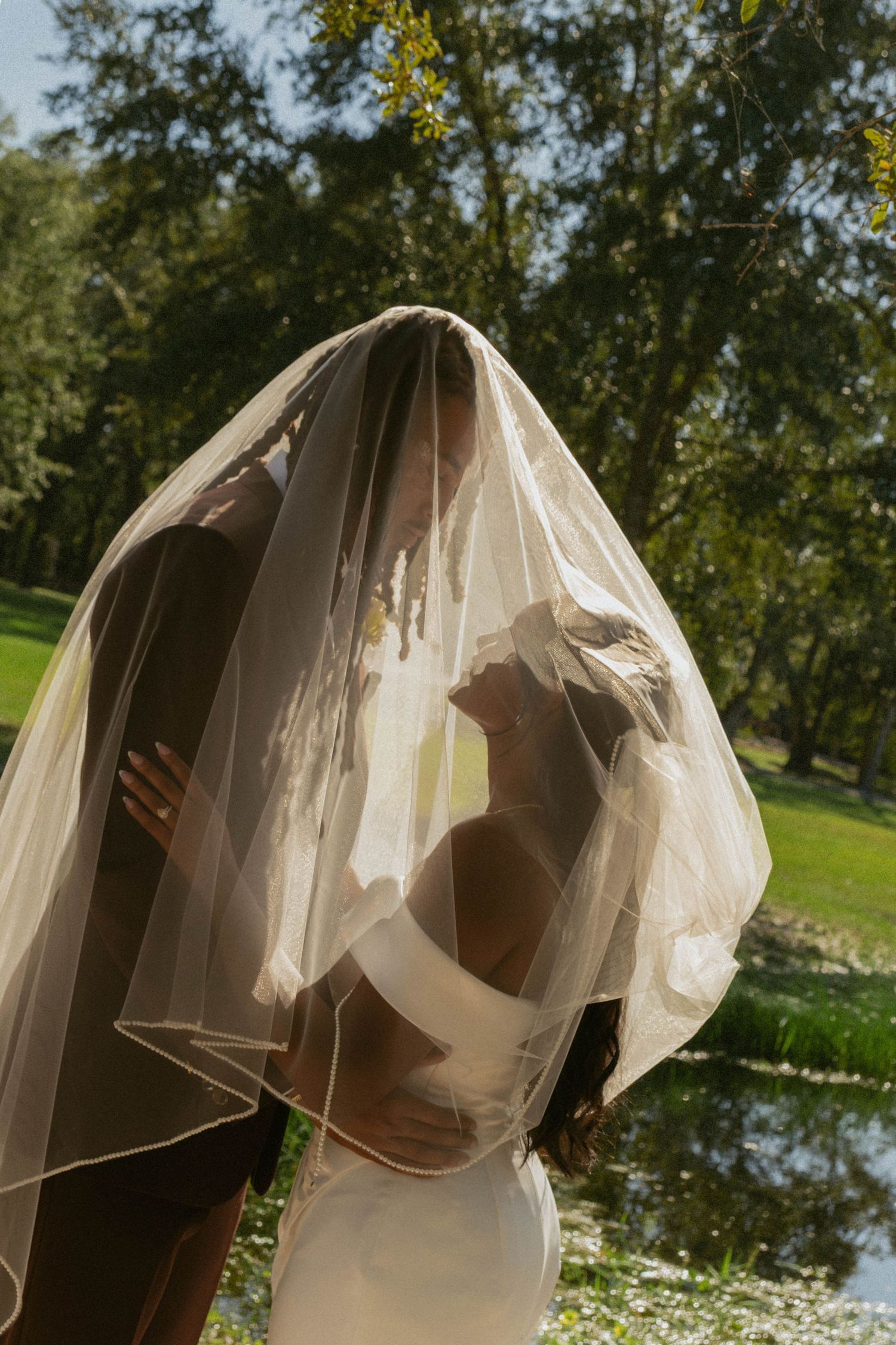 Elegant wedding photo with bride and groom under veil with trees and greenery in the background.