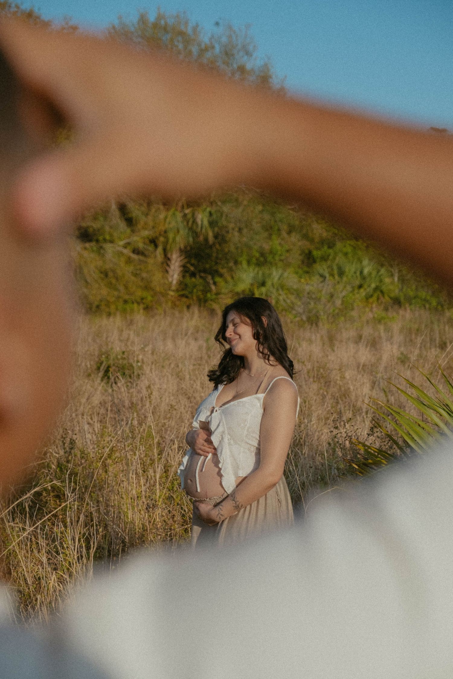 Woman in beige dress stands in golden field during sunset with blurred foreground elements creating dreamy atmosphere.