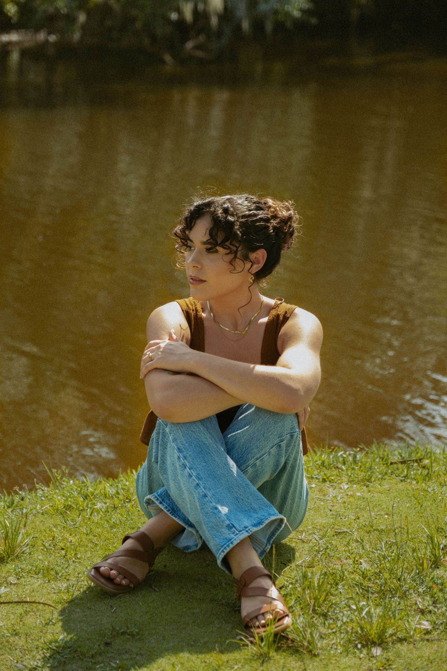 Person in casual wear sitting by lakeside on grass in warm sunlight.