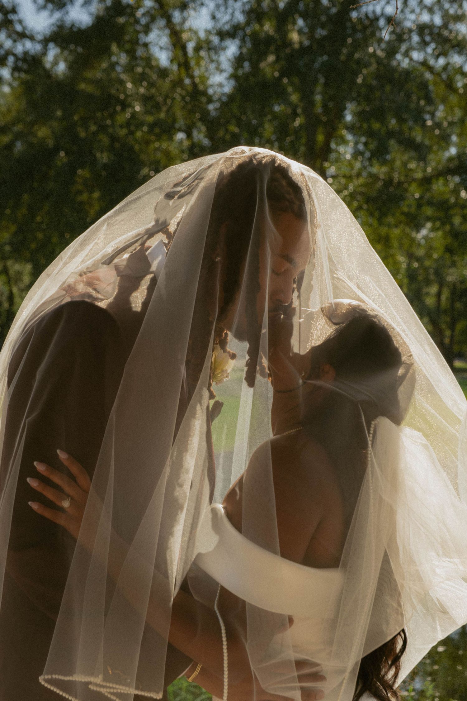 A romantic moment under a flowing white wedding veil against a sunlit background of trees.