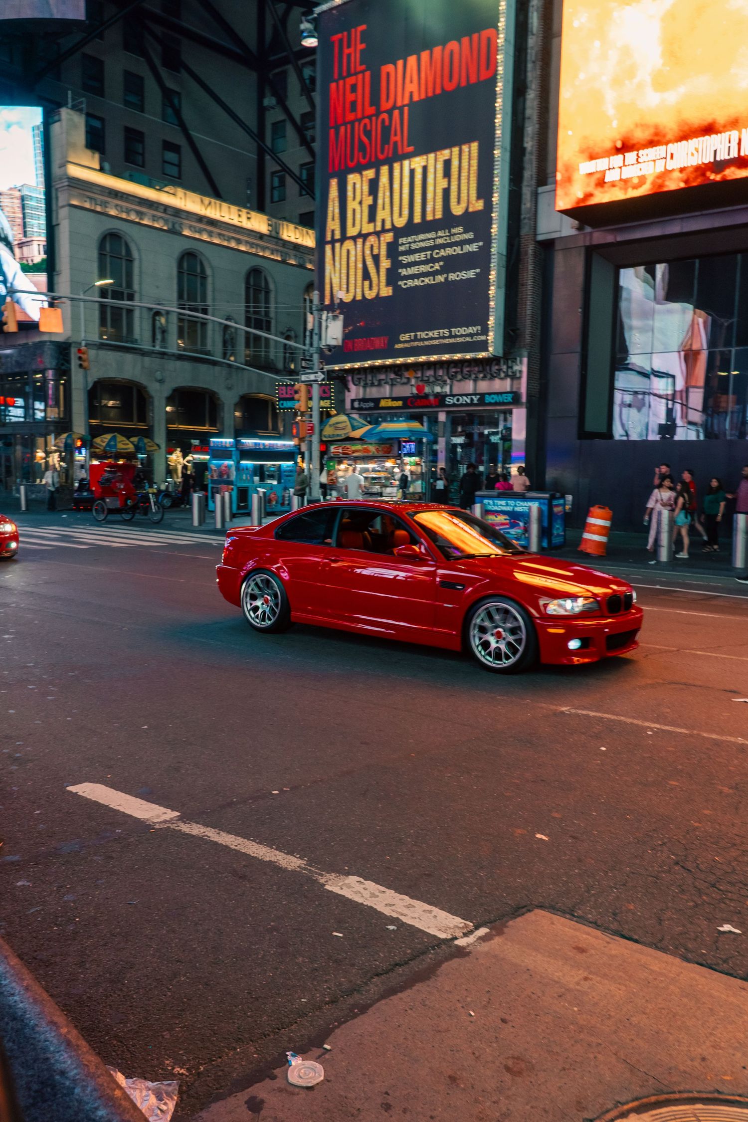 A red BMW M3 sports car drives through Times Square at night with bright theater billboards in the background.