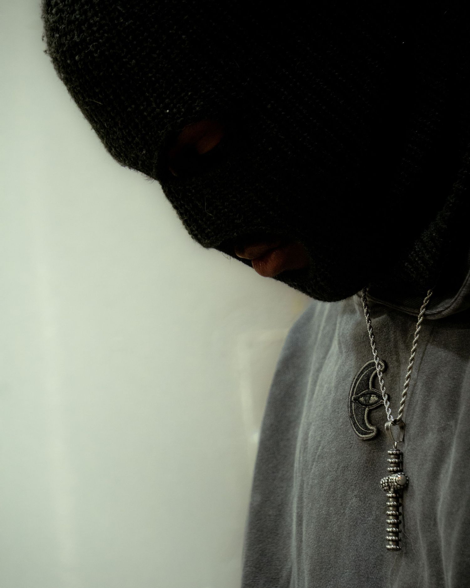 Silhouette of a person wearing a black ski mask and silver necklace against a light background.