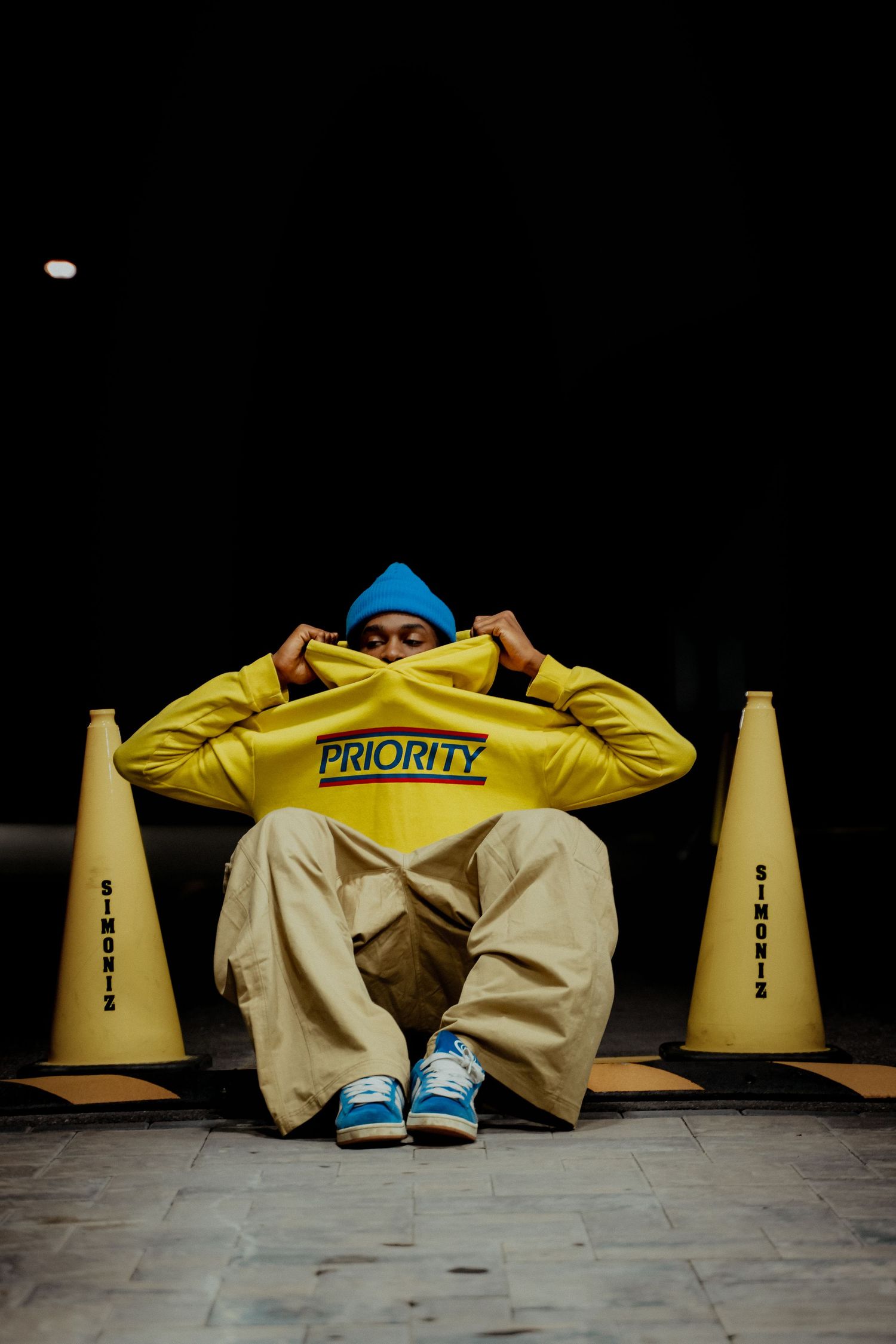 Person in yellow PRIORITY hoodie and khaki pants sitting between yellow traffic cones on floor.