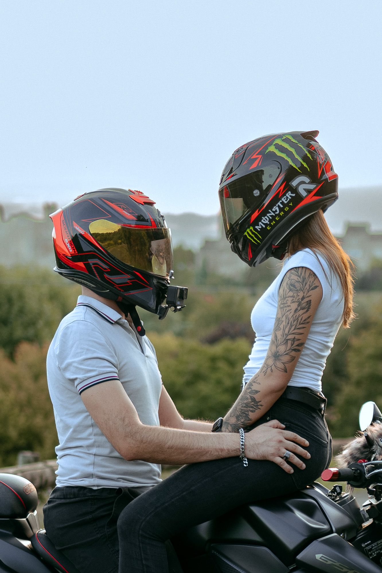 Two motorcyclists in black helmets and white shirts share an intimate moment while seated on a motorcycle.