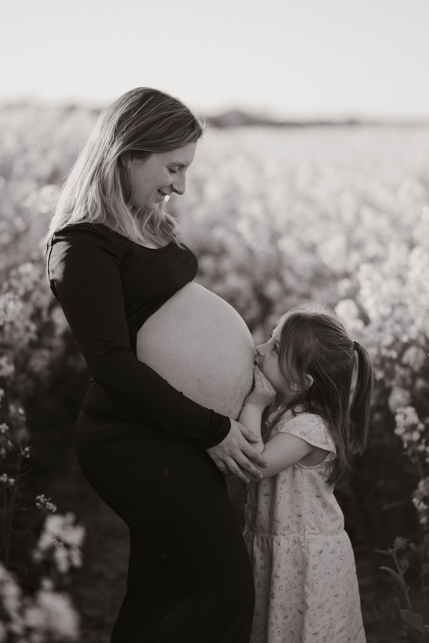 Black and white photo of a pregnant woman smiling as her young daughter kisses her baby bump in a flower field.