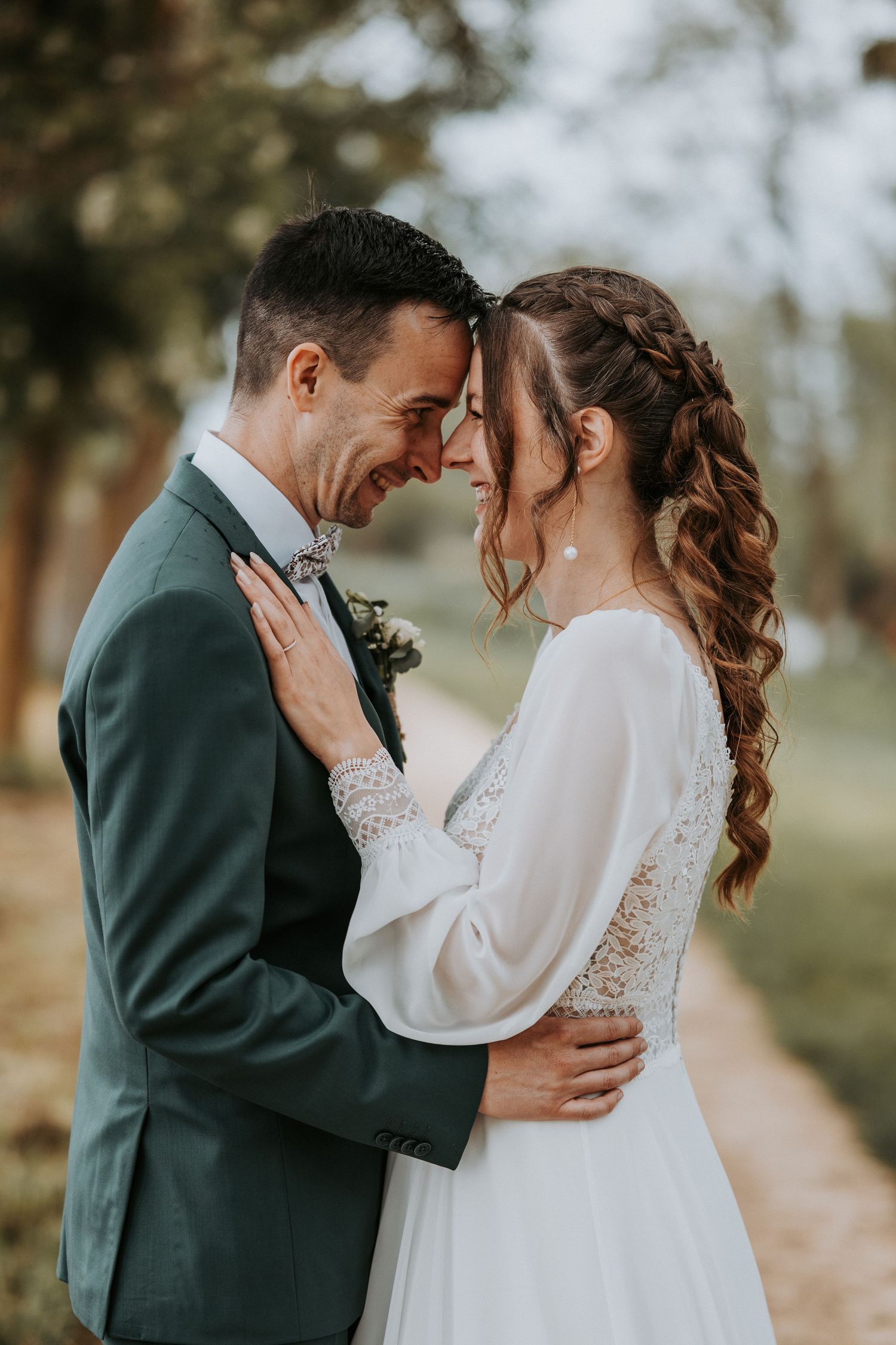 A bride and groom share a romantic forehead touch outdoors, she in a white lace gown, he in a green suit.