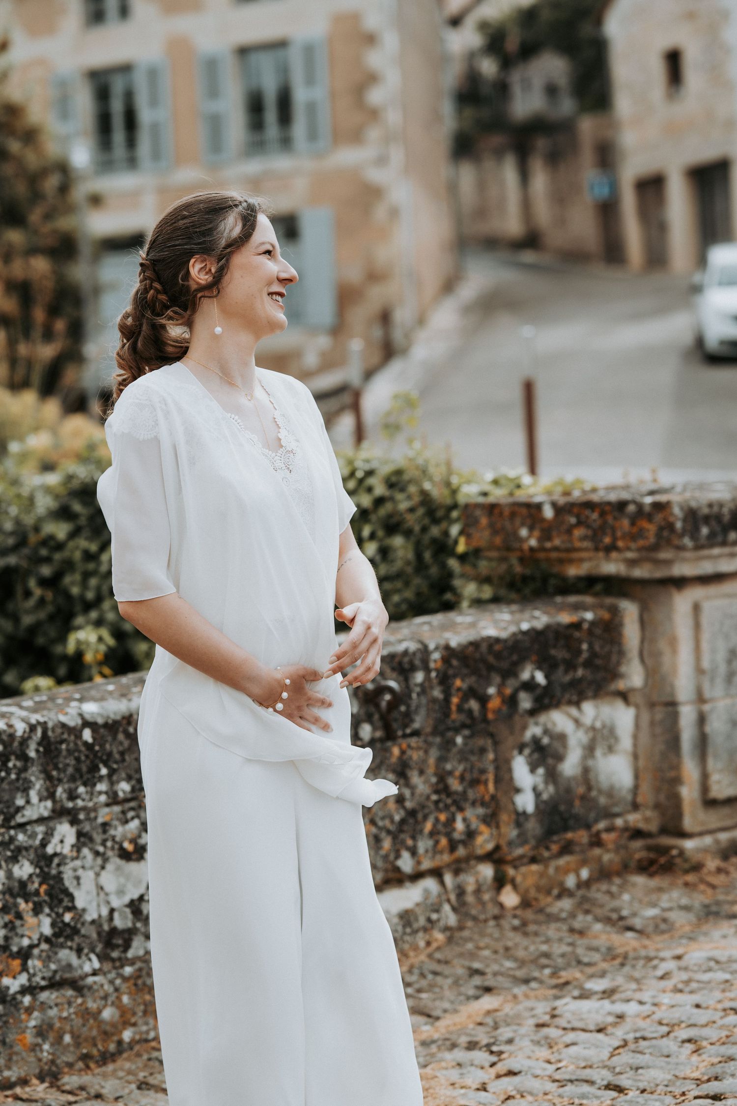 Smiling bride in white jumpsuit standing on cobblestone street in charming European village setting.