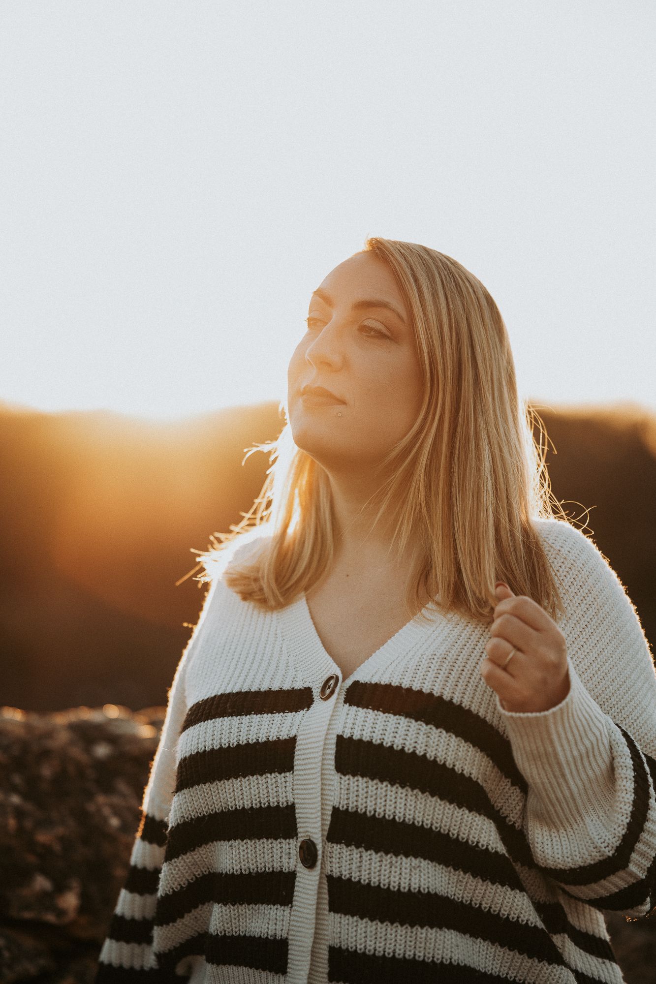 Blonde woman in striped knit cardigan poses outdoors during golden sunset, warm backlight illuminating her hair.