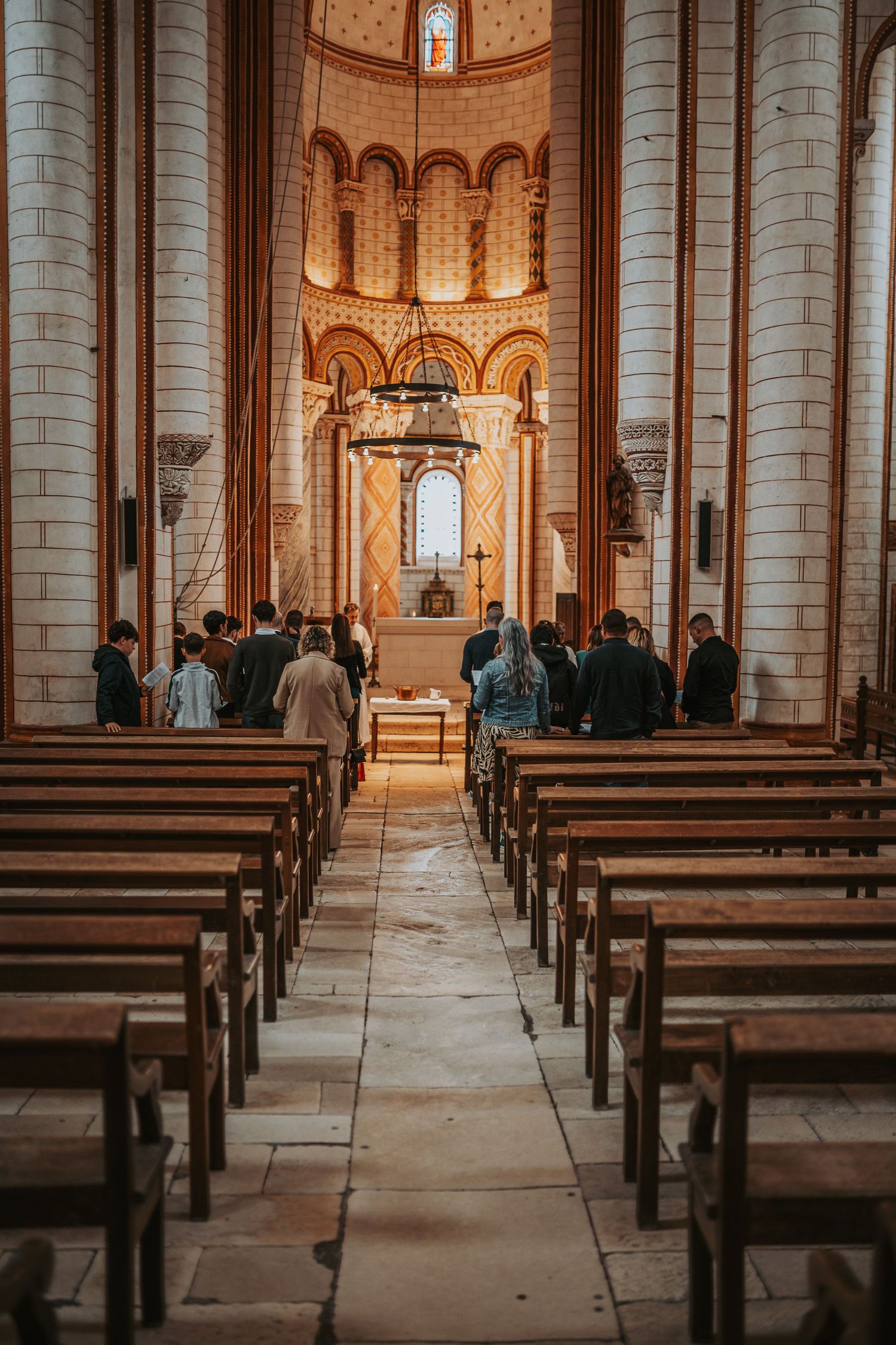 People gathered at the altar of a grand Romanesque church with wooden pews lining the stone-tiled nave.