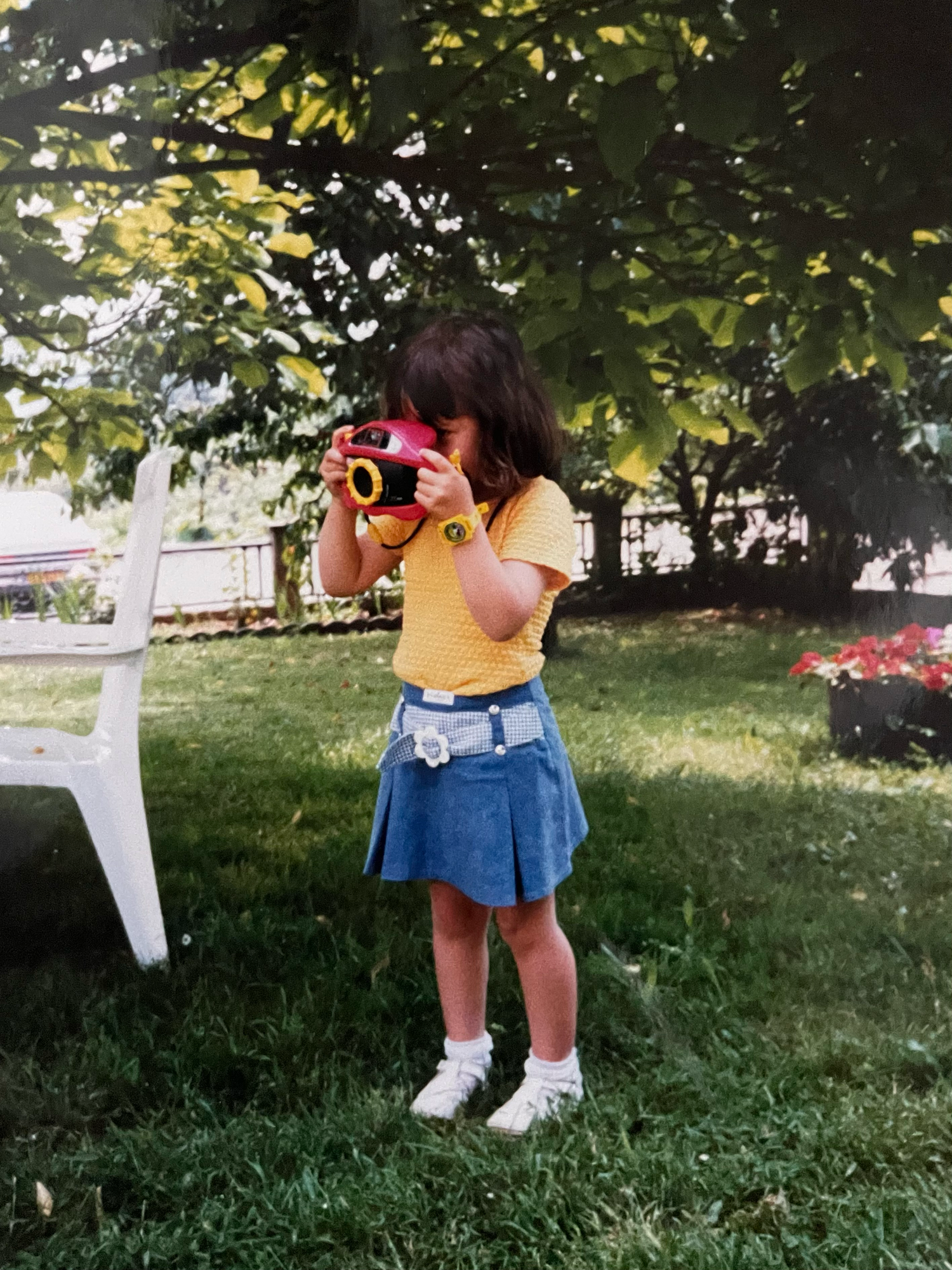Young girl in yellow shirt and denim skirt holds colorful toy camera outdoors on green lawn near trees.