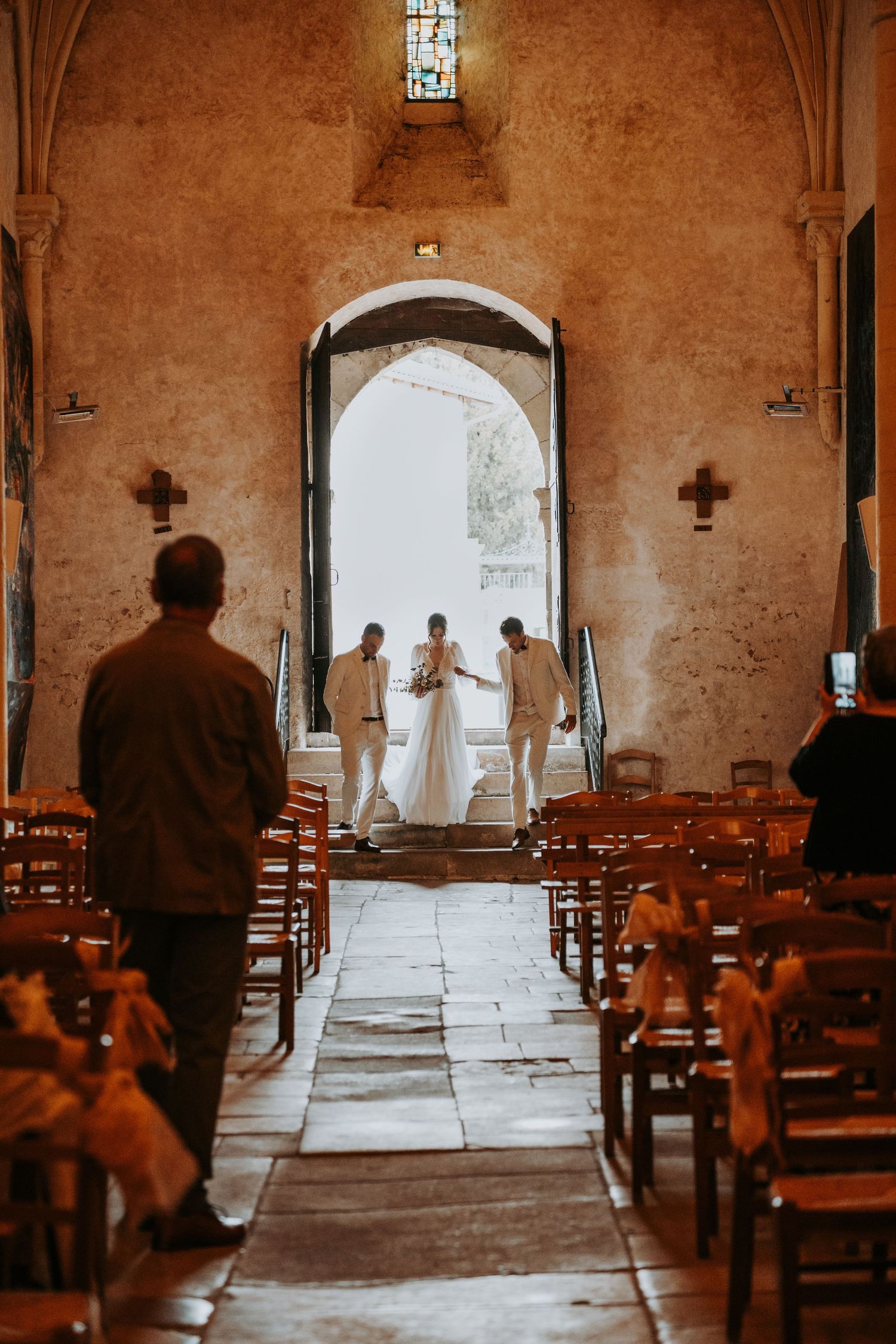Bride walking down the aisle of a rustic stone church with groomsmen, warm light flooding through grand entrance doors.