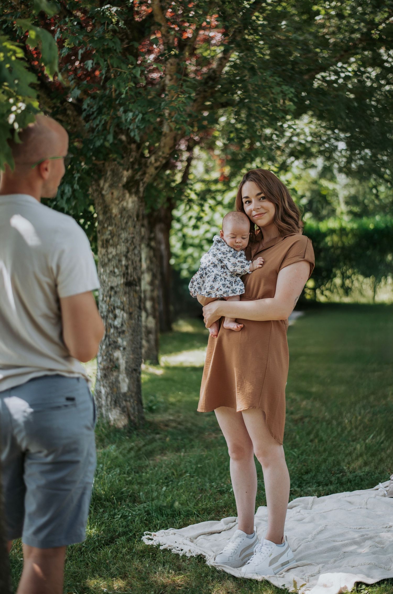 Family taking outdoor photos together on a white blanket under a tree in a park setting on a sunny day.