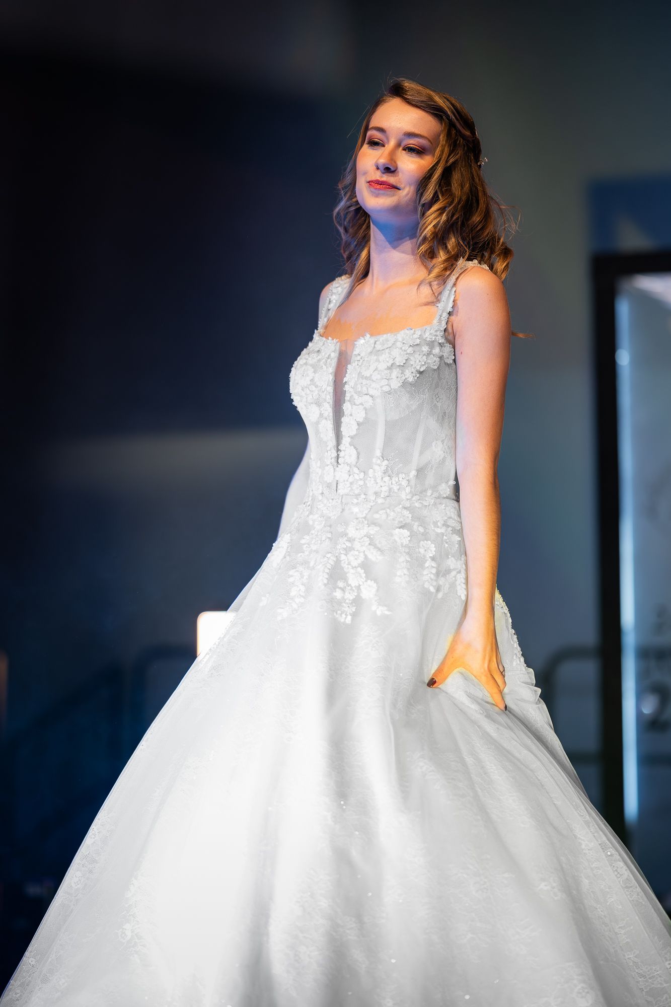 A model showcases a stunning white ballgown wedding dress with lace details and a full tulle skirt on a dark stage.